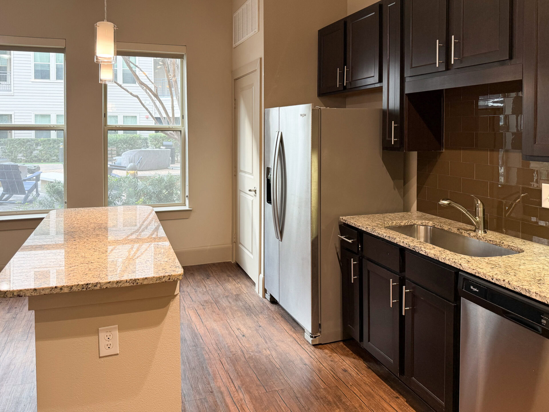 A modern kitchen features dark cabinetry and a light granite countertop. The stainless steel refrigerator stands next to a sink with a sleek faucet. A window provides natural light, revealing a view of an outdoor area with seating. The flooring is a warm wood tone, enhancing the contemporary feel of the space.