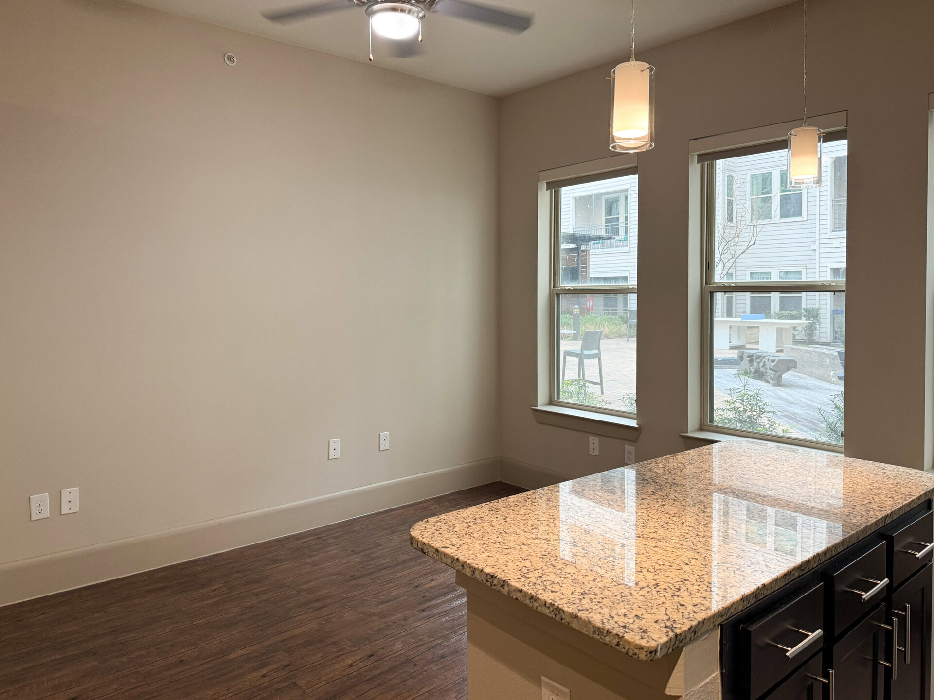 A spacious kitchen area features a large granite countertop with a smooth, polished surface. Two large windows allow natural light to flood the space, offering a view of an outdoor area with greenery and seating. The walls are painted a neutral color, and the flooring is dark wood. Two pendant lights hang above the countertop, adding a modern touch. Electrical outlets are visible on the wall, enhancing the functionality of the space.