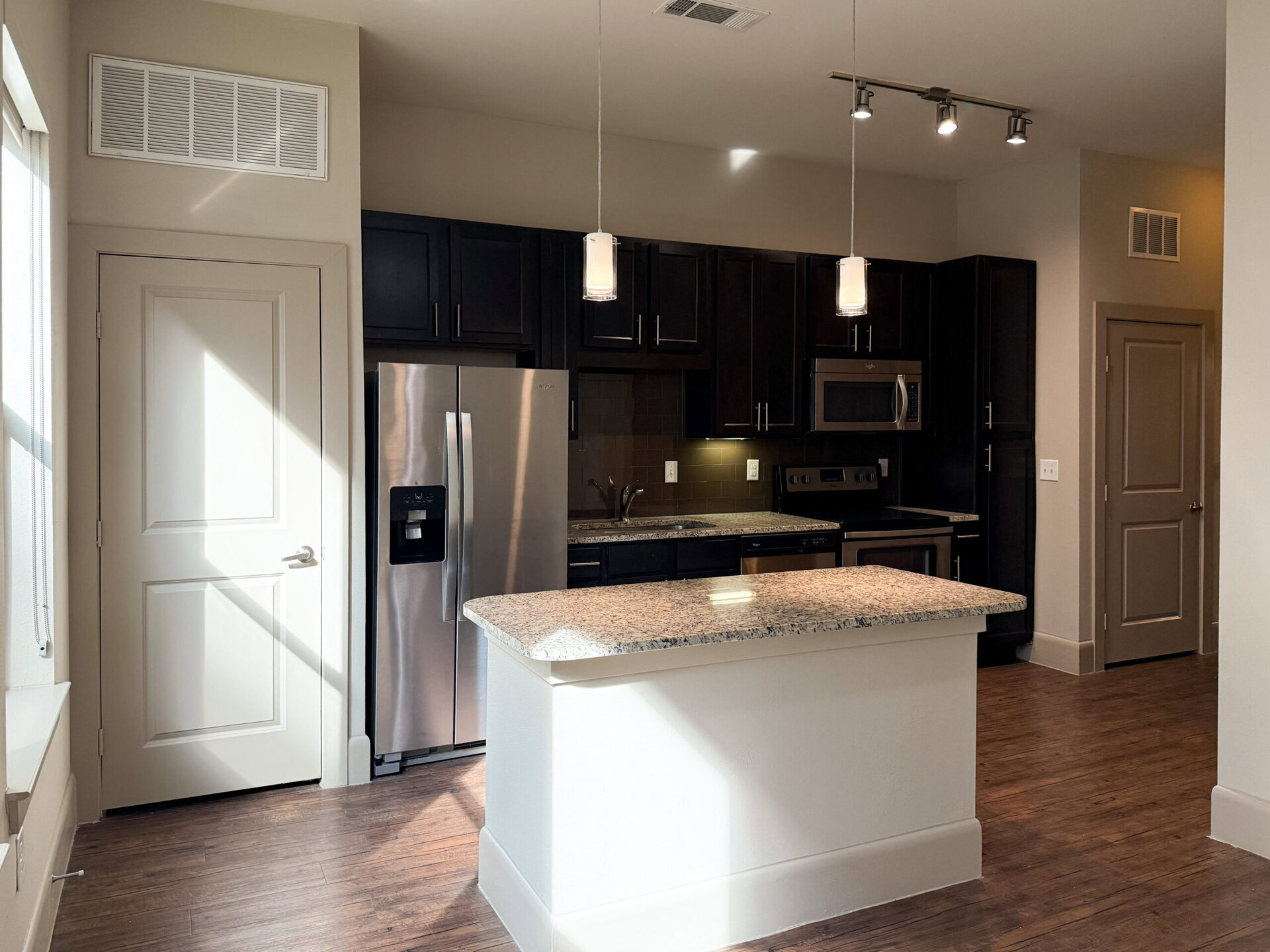 A modern kitchen features dark wood cabinetry and stainless steel appliances, including a refrigerator, microwave, and oven. The countertop is made of light-colored granite, providing a contrast to the dark cabinets. Two pendant lights hang above the countertop, illuminating the space. A door is visible on the left, and natural light streams in from a window, casting shadows on the wall. The flooring is a warm wood tone, enhancing the contemporary feel of the kitchen.