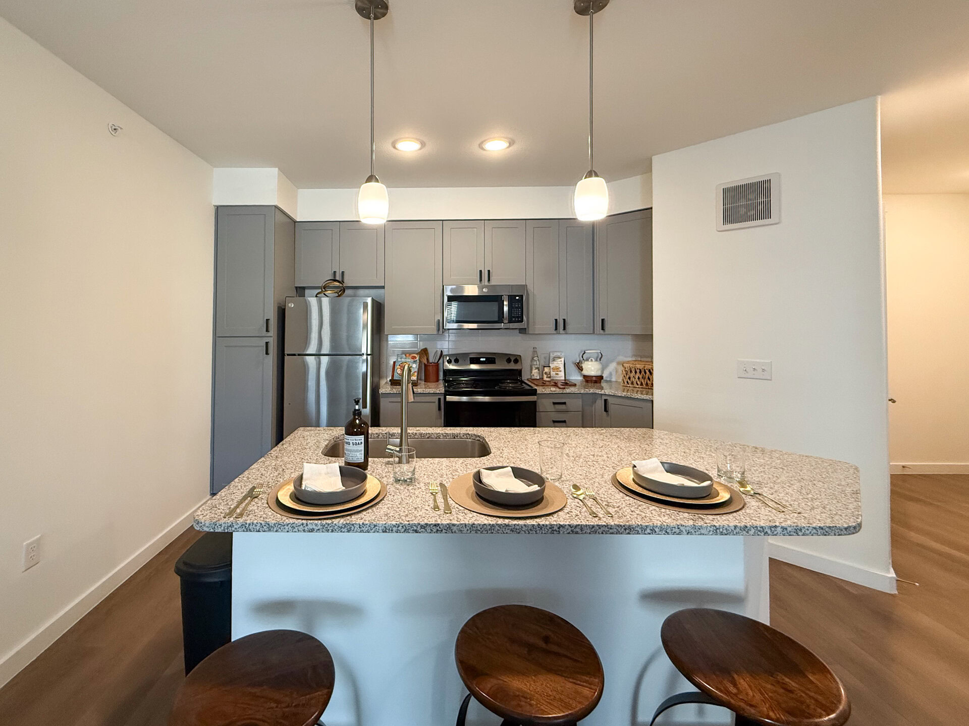 A modern San Antonio apartment kitchen features gray cabinetry and stainless steel appliances, including a refrigerator and stove. The countertop is made of speckled granite with modern light fixtures above. The flooring is light wood, and overall, the unit has plenty of natural sunlight.