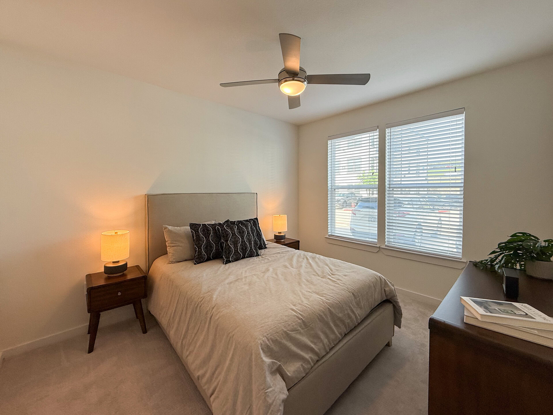 A cozy San Antonio apartment bedroom features a ceiling fan with a light fixture hangs above, and two large windows with blinds allow natural light to fill the room. The room has a bright, inviting atmosphere.