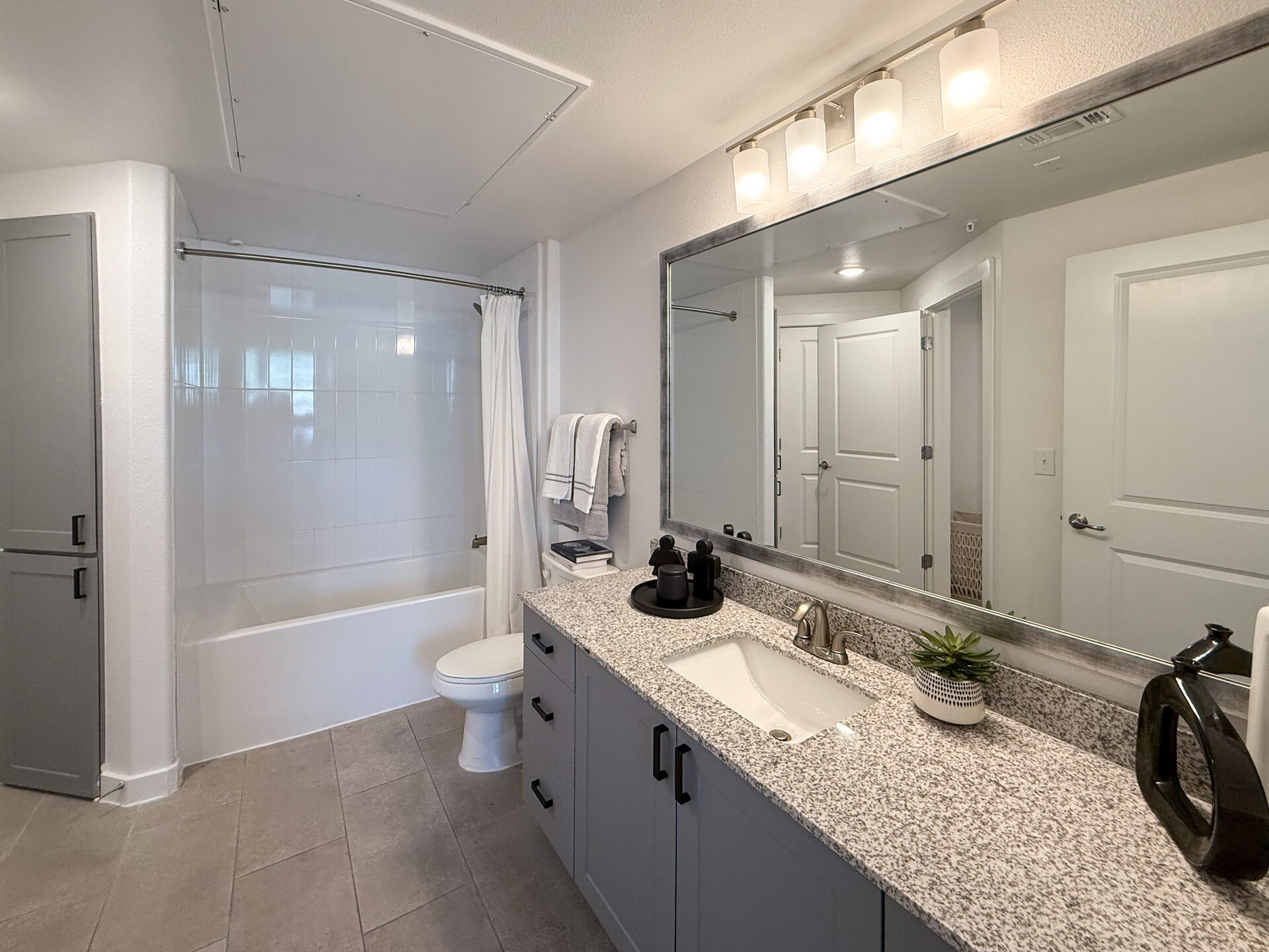 A modern San Antonio apartment bathroom features a white bathtub with a shower curtain, a toilet, and a sink vanity with a granite countertop. The vanity has a sleek faucet. Above the sink, a large mirror is framed in metal, reflecting the light from three glass light fixtures. The walls are painted in a light color, and there are gray cabinets for storage. The flooring is tiled in a neutral shade.