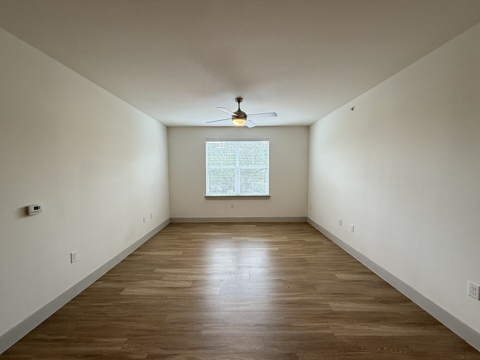 A spacious, empty room with light-colored walls and a wooden floor. A ceiling fan with a light fixture is mounted in the center. On one side, there is a window with blinds, allowing natural light to enter. The room has minimal furnishings and is ready for decoration or furniture placement.