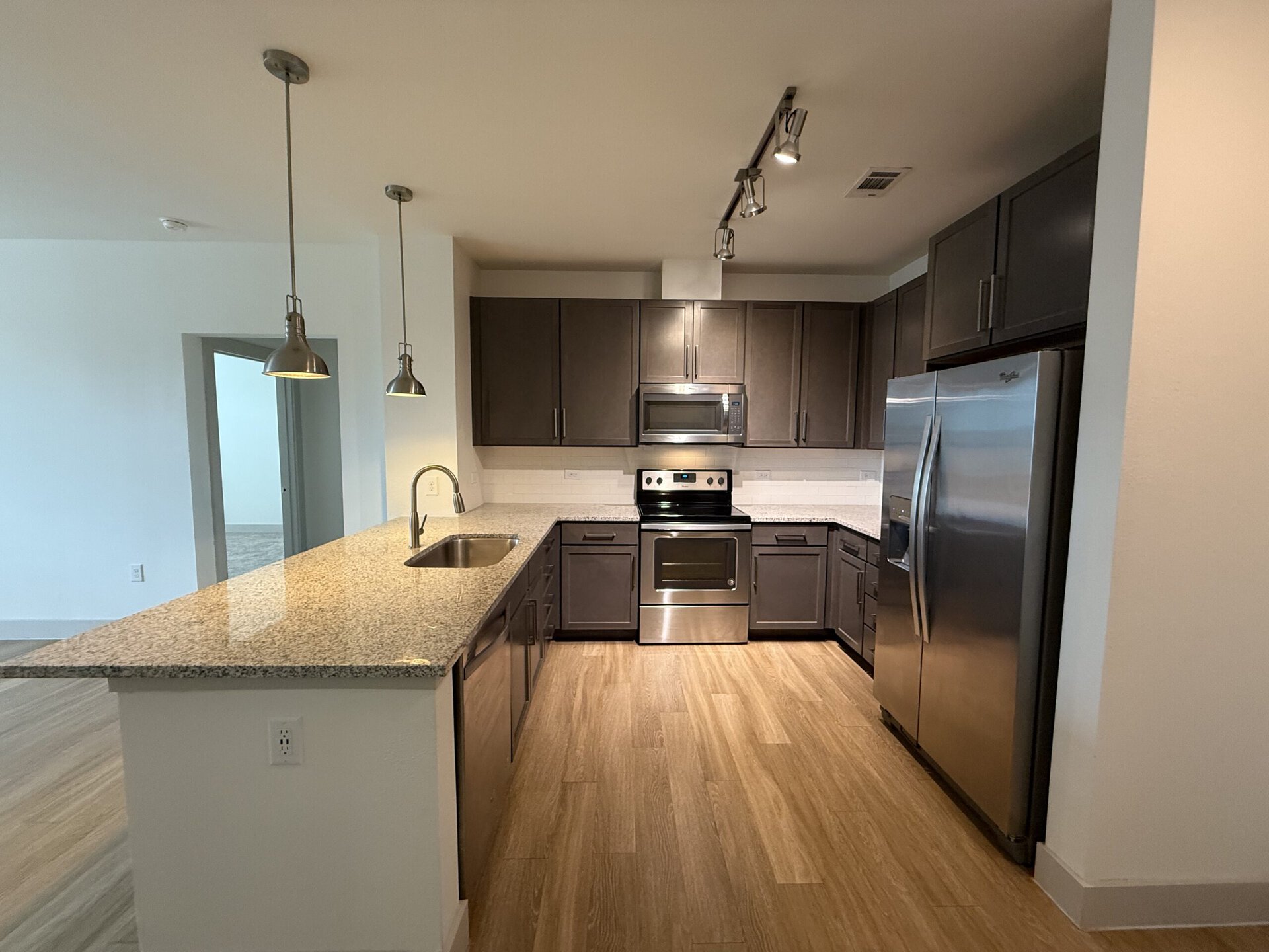 A modern kitchen featuring dark wood cabinets and a light granite countertop. The kitchen includes stainless steel appliances, including an oven, microwave, and refrigerator. Pendant lights hang above the countertop, which has a sink integrated into it. The flooring is a light wood laminate, and the walls are painted a neutral color. A doorway is visible in the background, leading to another room.