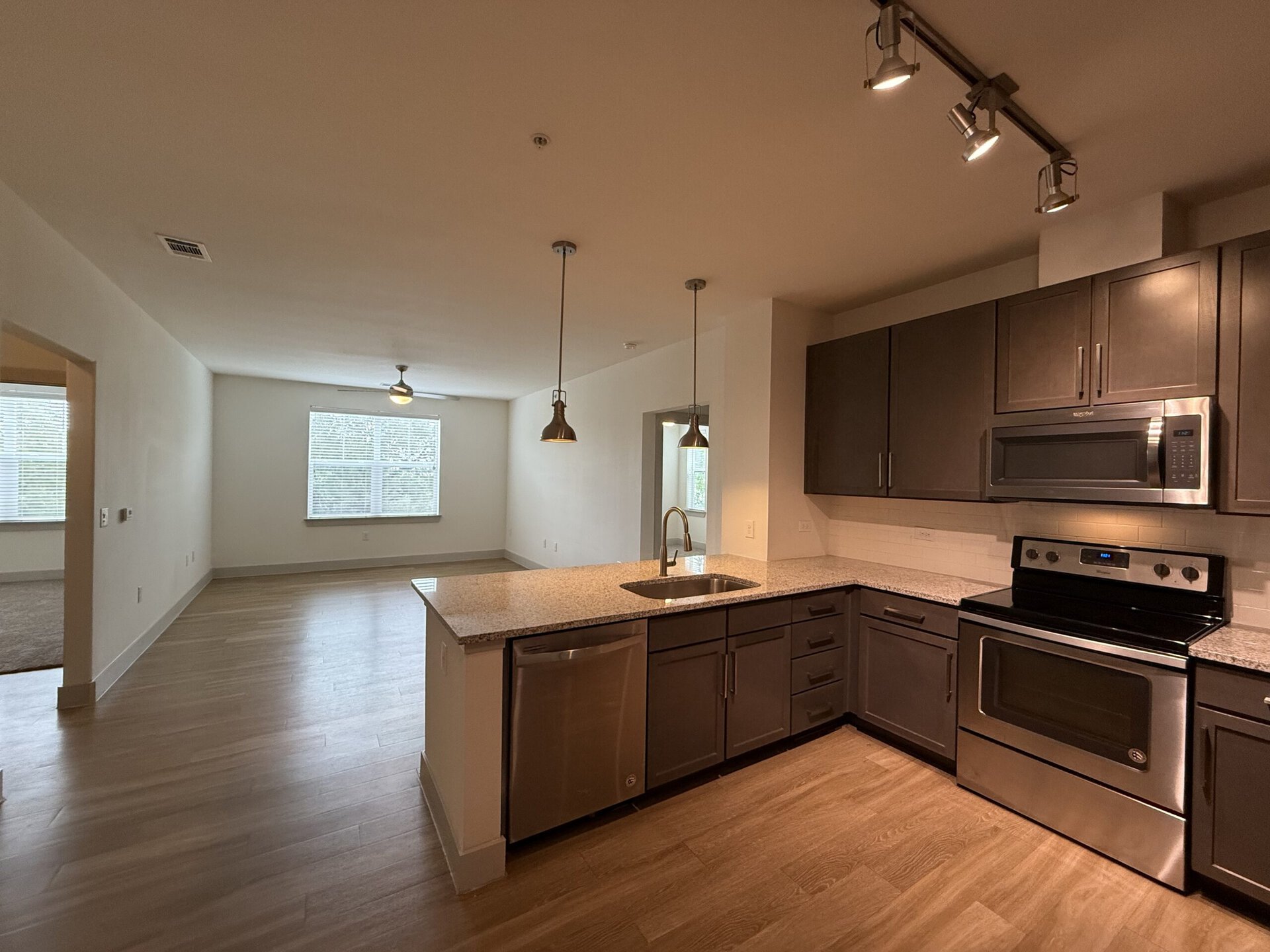 A modern kitchen area features dark cabinetry and stainless steel appliances, including a microwave and an oven. The countertop is made of light-colored granite, and a stainless steel sink is installed with a sleek faucet. Two pendant lights hang above the counter. In the background, a spacious living area is visible, with large windows allowing natural light to enter, and light wood flooring throughout. The walls are painted in a neutral tone, enhancing the contemporary feel of the space.