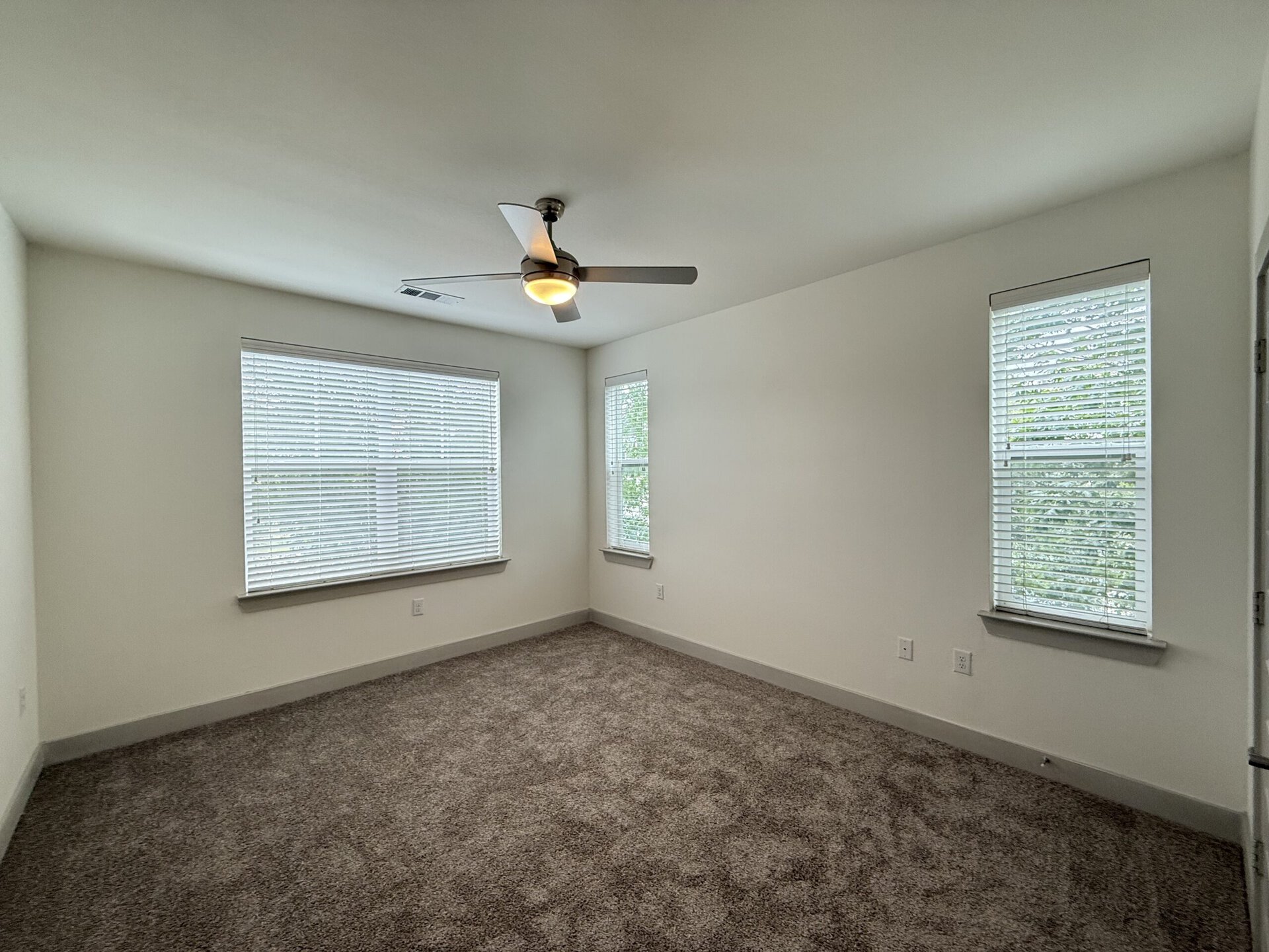 A spacious room features light-colored walls and plush, beige carpeting. Two windows with white blinds allow natural light to enter, creating a bright atmosphere. A modern ceiling fan with a light fixture is mounted in the center of the ceiling. The room is uncluttered, providing a blank canvas for furniture arrangement.