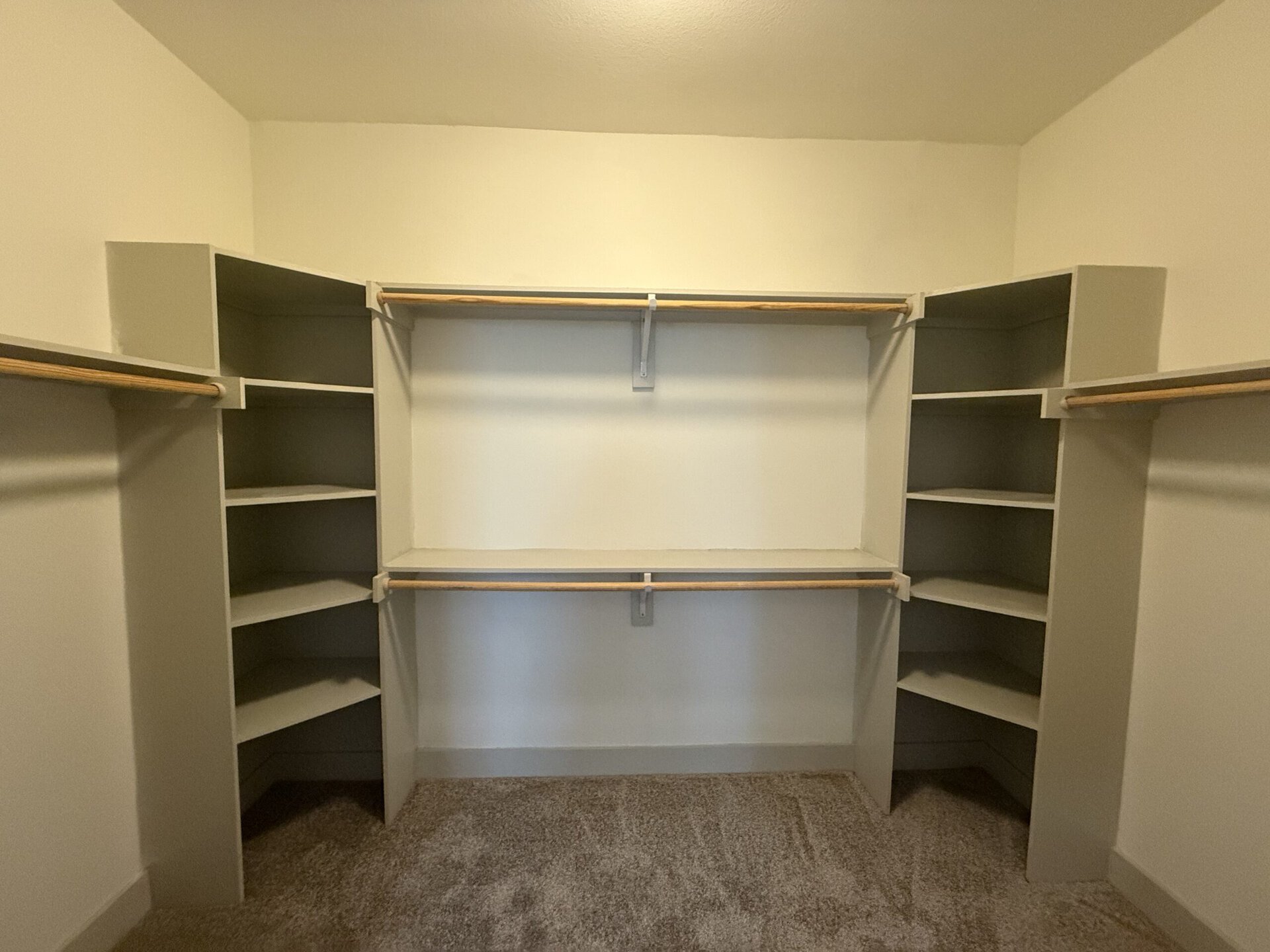 A spacious closet features built-in shelving and hanging rods. The walls are painted a light color, and the shelves are arranged in a U-shape with a central section for additional storage. The flooring is carpeted, providing a soft surface. The design allows for ample organization and accessibility.