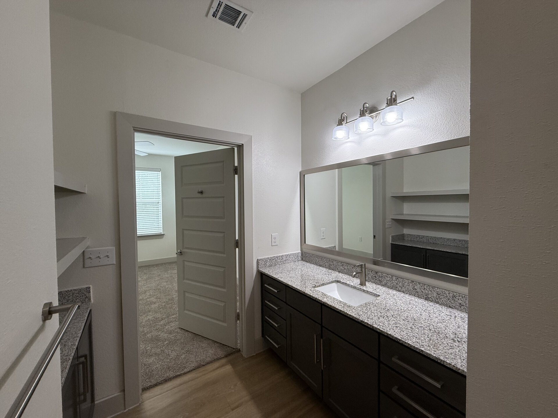 A modern bathroom features a sleek vanity with a white sink and a granite countertop. Above the sink, there is a large mirror framed in metal, accompanied by three light fixtures. To the left, a door leads to another room, while a shelf is mounted on the wall nearby. The walls are painted in a light color, and the flooring appears to be a warm wood tone. Natural light enters from a window in the adjacent room, which has blinds.