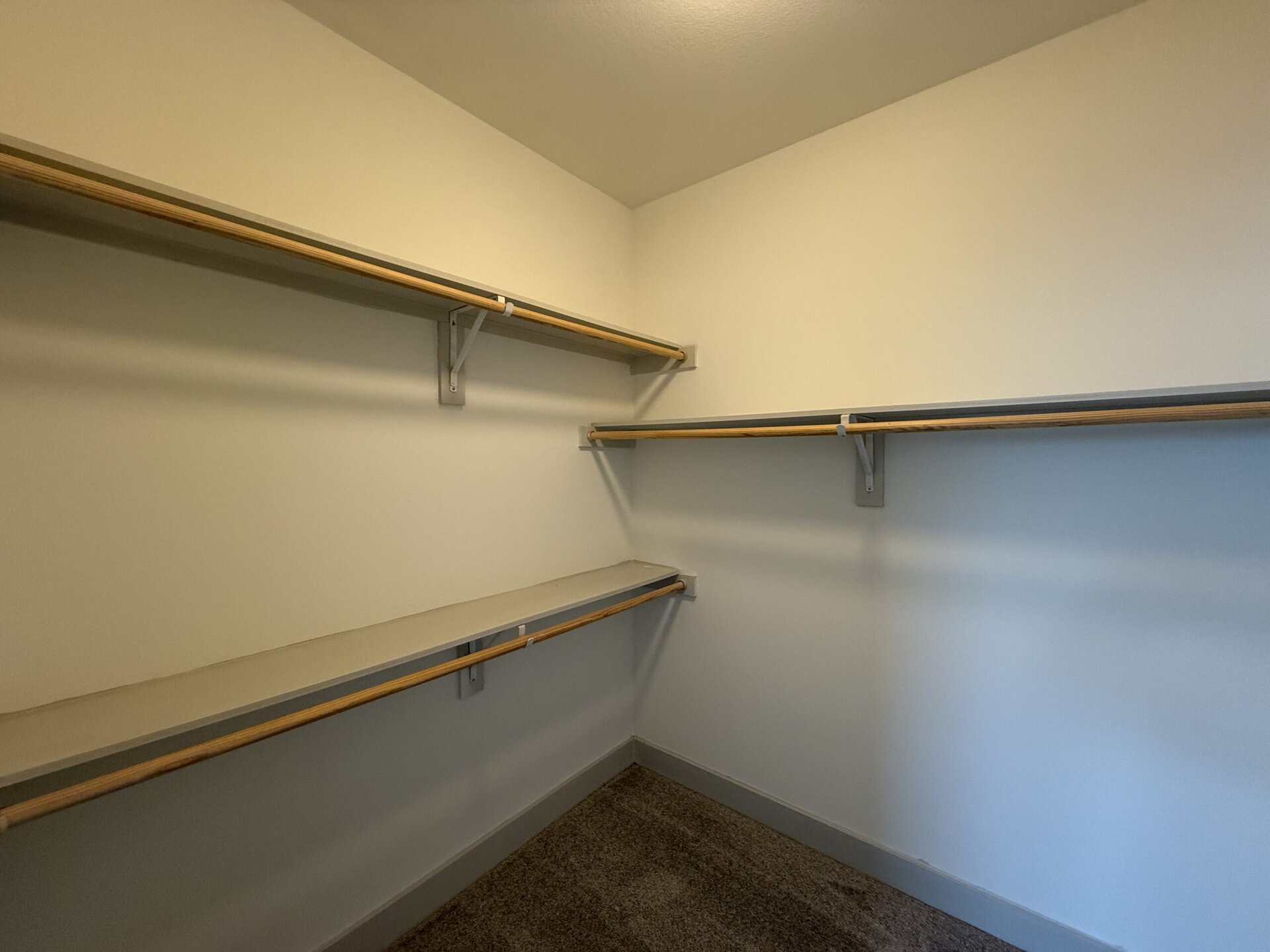 A spacious closet features two wooden shelves mounted on the walls, with a lower shelf running along one side. The walls are painted a light color, and the floor is carpeted. The overall design is simple and functional, providing ample storage space.