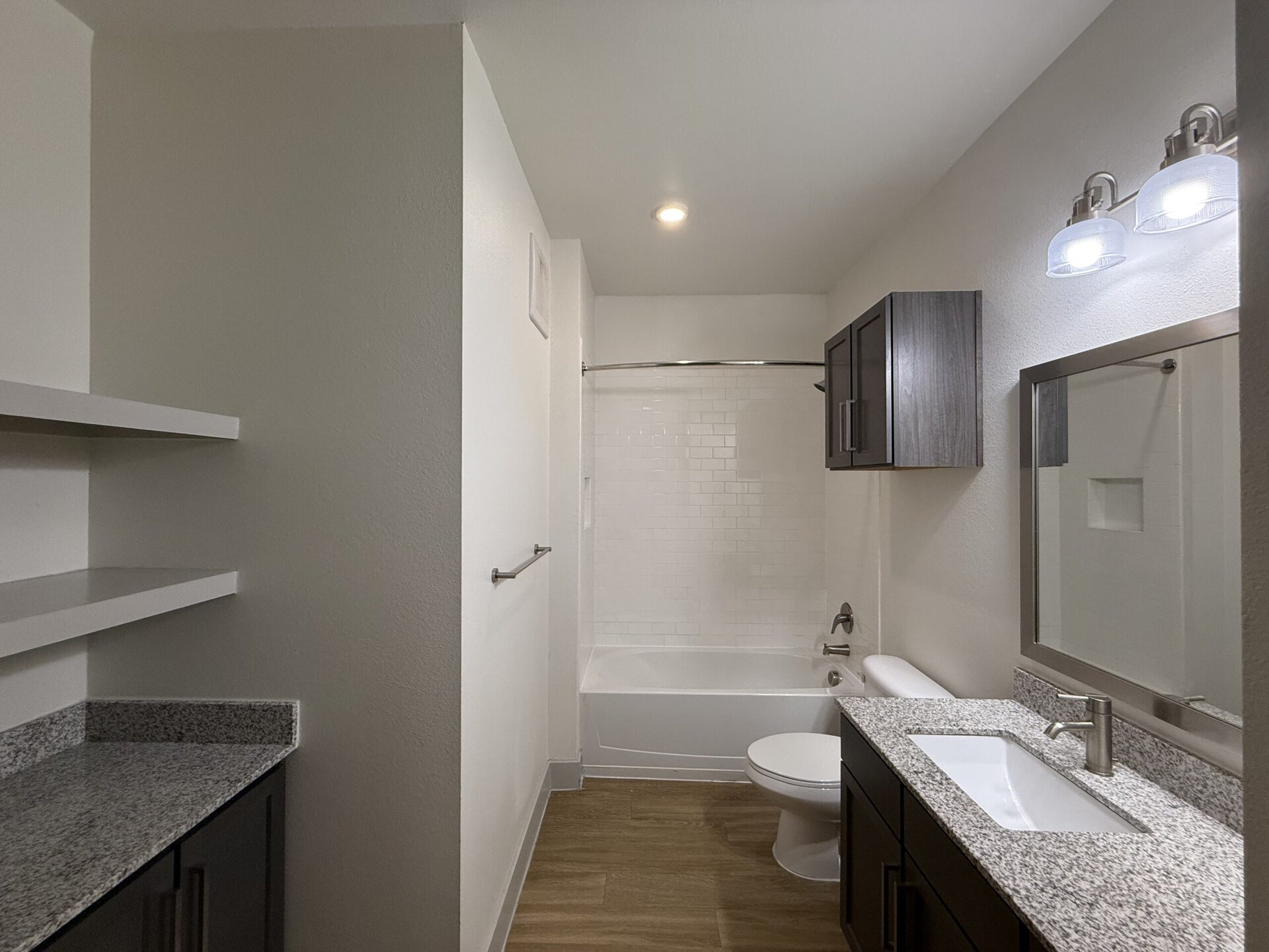 A modern bathroom featuring a bathtub with a shower, surrounded by white subway tiles. The space includes a dark wood vanity with a granite countertop and a rectangular sink. Above the vanity, there is a large mirror framed in silver, complemented by two stylish light fixtures. To the left, open shelving provides additional storage, while a towel bar is mounted on the wall. The flooring is a light wood finish, adding warmth to the overall design.