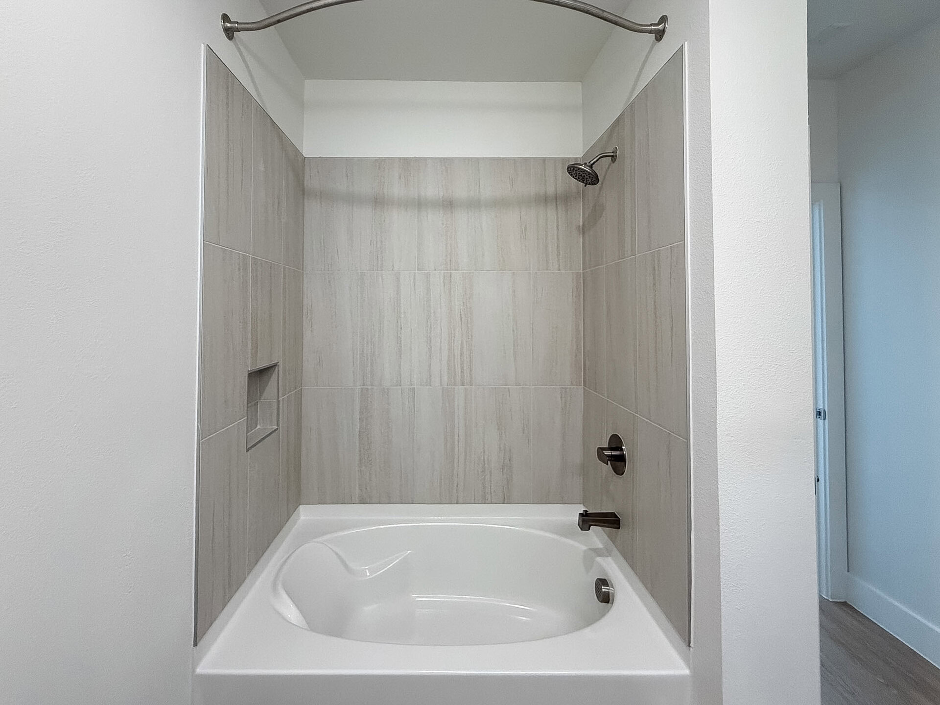 A modern San Antonio apartment bathroom tub shower features the interior having light-colored, textured tiles, creating a clean and spacious look. A small shelf is integrated into the wall for convenience. The surrounding walls are painted in a soft white, complementing the overall aesthetic.