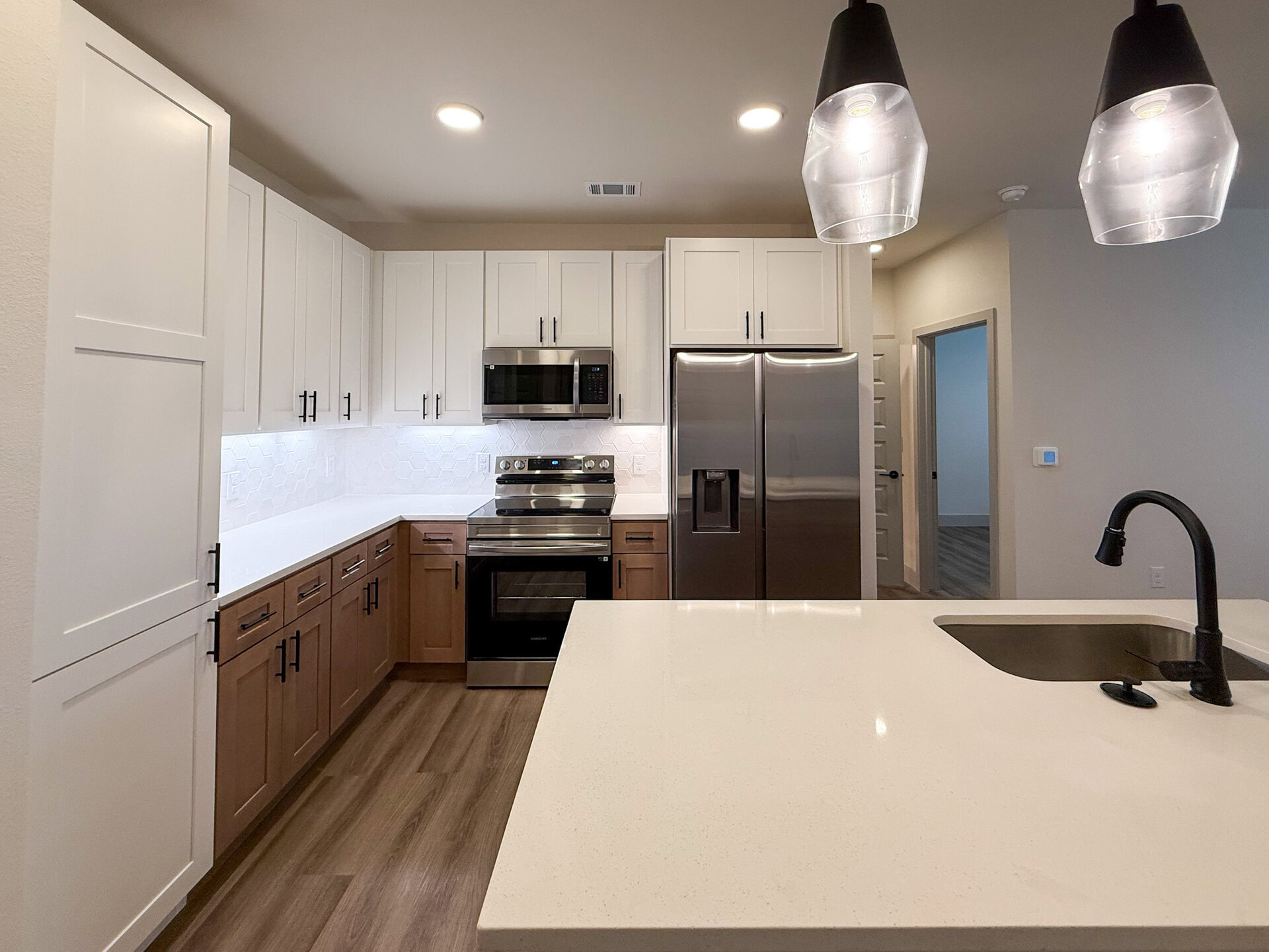 A modern Northwest San Antonio apartment kitchen featuring a combination of light and dark cabinetry. The upper cabinets are white with black handles, while the lower cabinets are a warm wood tone. A stainless steel double door refrigerator and microwave are positioned next to a sleek oven with a glass cooktop. The backsplash consists of white hexagonal tiles, and the countertop is a smooth, white surface. A matte black faucet is installed over a sink integrated into the island. Two pendant lights hang above the kitchen island, illuminating the space. The flooring is a light wood laminate.