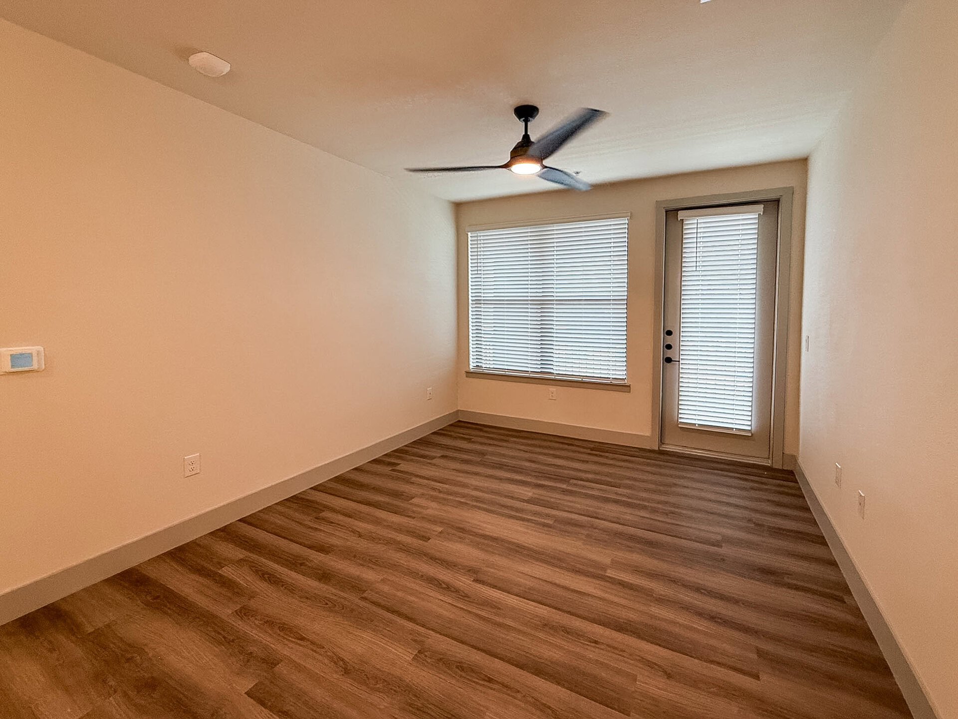 A spacious Northwest San Antonio apartment living room features light-colored walls and a wood-like floor. A ceiling fan is mounted in the center, providing a modern touch. On one side, there is a large window with horizontal blinds, allowing natural light to enter. Adjacent to the window, a glass door with blinds leads outside, enhancing the room's accessibility. The overall atmosphere is bright and inviting, suitable for various uses.
