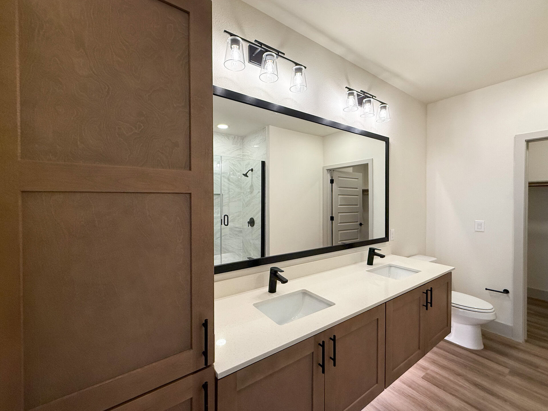 A modern San Antonio apartment bathroom features a single vanity with a light-colored countertop. The cabinetry is a warm wood tone with black handles. Above the vanity, a large rectangular mirror with a black frame reflects the space. A glass shower enclosure with marble-like tiles. The overall design is sleek and modern, with neutral colors and clean lines.