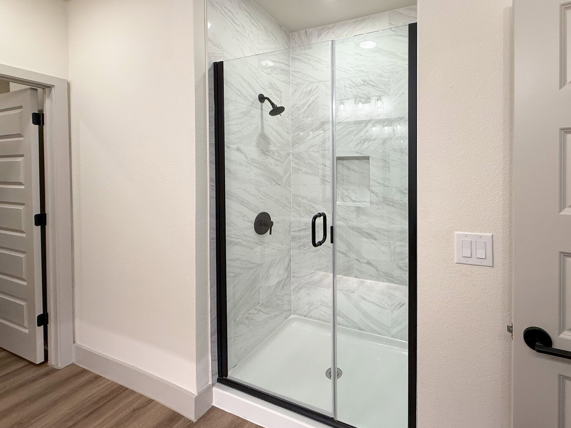 A modern luxe Northwest San Antonio apartment standing shower features a glass door with a black frame. Inside, the walls are covered with white marble tiles arranged in a herringbone pattern. A rainfall showerhead is mounted on the wall, and there is a built-in niche for toiletries. The floor is a smooth, white surface. The overall aesthetic is clean and modern luxe.