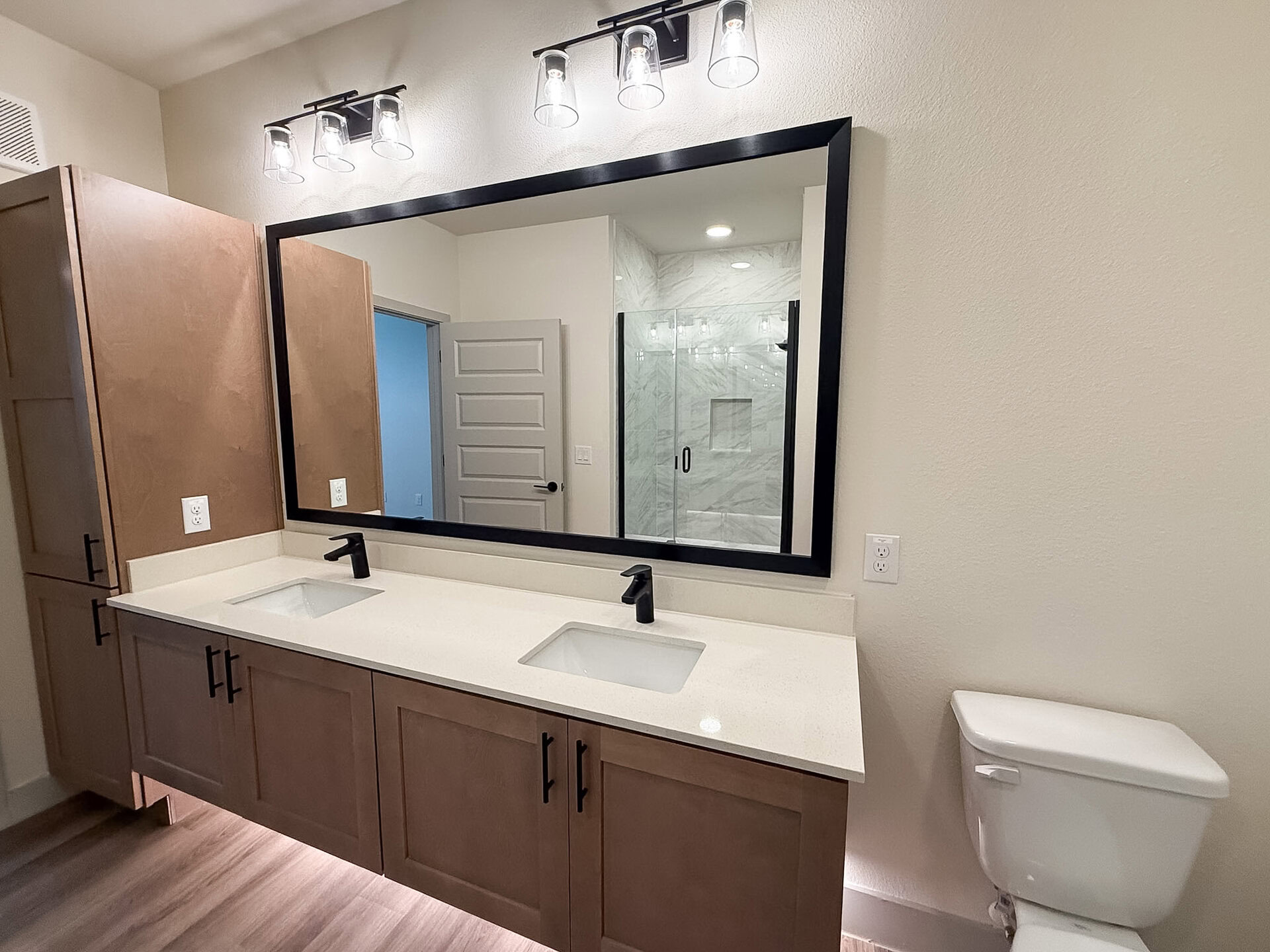 A modern San Antonio apartment bathroom features a double vanity with a light-colored countertop and two square sinks. The cabinetry is a warm wood tone with black handles. Above the vanity, a large rectangular mirror with a black frame reflects the space. A glass shower enclosure with marble-like tiles. The overall design is sleek and modern, with neutral colors and clean lines.