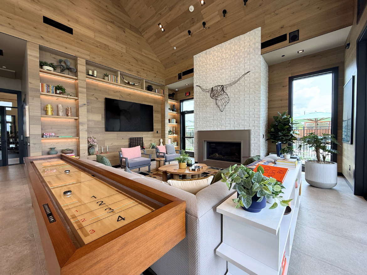 A modern living room features a high, wooden ceiling and a large wall-mounted TV. The space is decorated with light wood paneling and built-in shelves displaying books and decorative items. A stylish fireplace is situated in the center, flanked by two comfortable armchairs with pink cushions. A shuffleboard table made of wood is prominently placed in the foreground. Various plants are arranged throughout the room, adding a touch of greenery. Large windows allow natural light to fill the space, with views of an outdoor area visible through the glass.