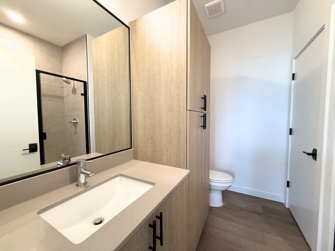 A modern bathroom features a light wood cabinetry with black handles, a sleek countertop with an integrated sink, and a large mirror above. To the left, there is a glass shower enclosure with tiled walls. A white toilet is positioned against the wall, and a door leads out of the space. The flooring is a light wood finish, contributing to the contemporary aesthetic.