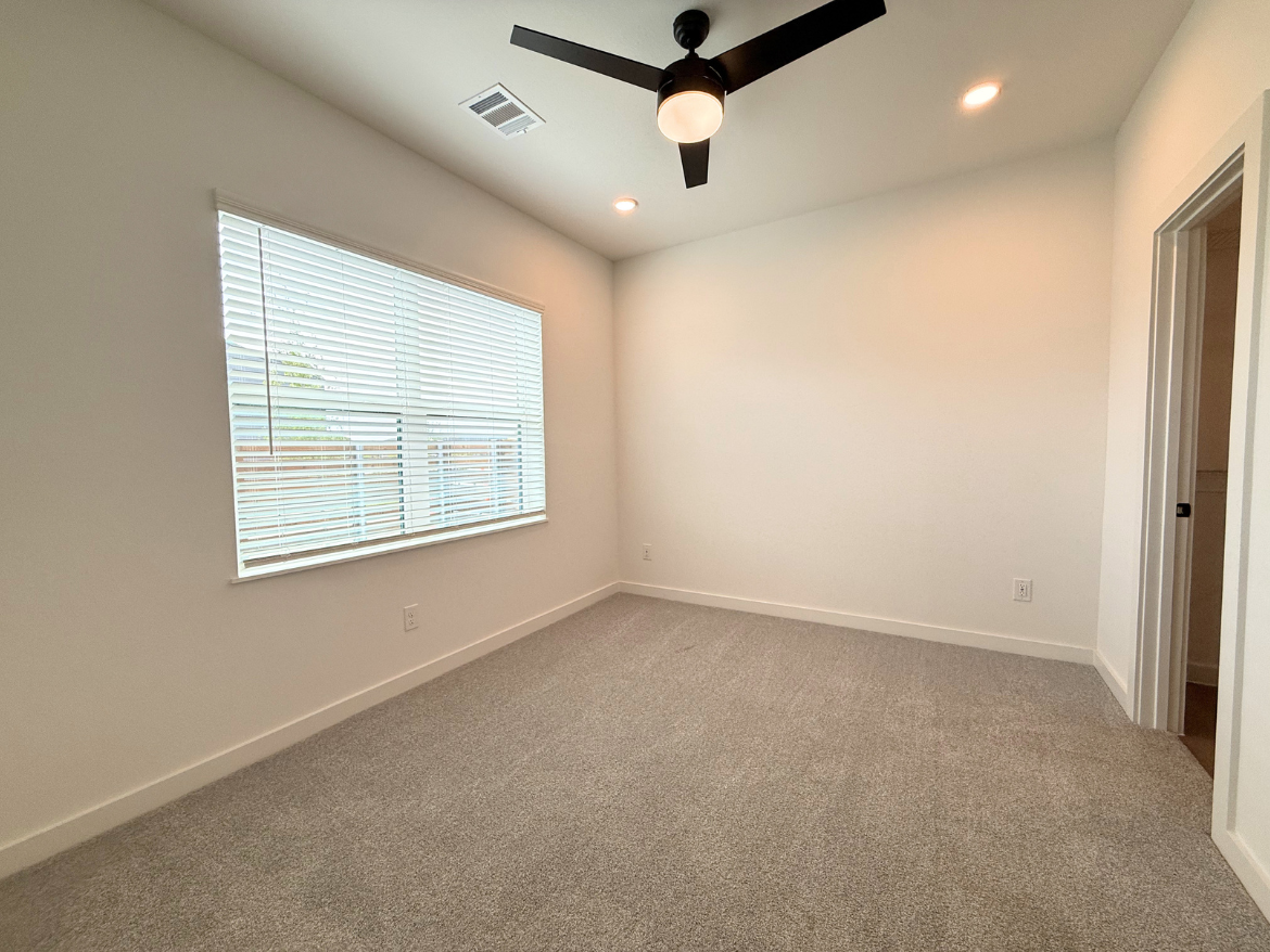 A spacious room with light gray carpet and white walls. A large window with horizontal blinds allows natural light to enter, illuminating the space. A modern ceiling fan with a light fixture hangs from the ceiling. On the right side, there is a doorway leading to another room or hallway. The overall aesthetic is clean and minimalistic.