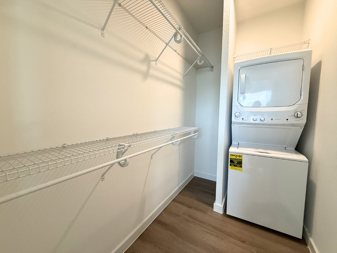 A compact laundry area features a stacked washer and dryer unit in white, positioned against a wall. To the left, there are two wire shelving units mounted above a long wire shelf, providing storage space. The walls are painted in a light color, and the flooring is a light wood laminate. The overall space is bright and organized, ideal for laundry tasks.