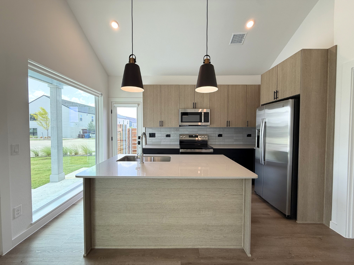 A modern kitchen features light wood cabinetry and a large island with a white countertop. Two pendant lights hang above the island. Stainless steel appliances, including a microwave and oven, are integrated into the cabinetry. A large window allows natural light to fill the space, showcasing a view of the outdoors with green grass and a fence. The flooring is a warm wood tone, complementing the overall design.