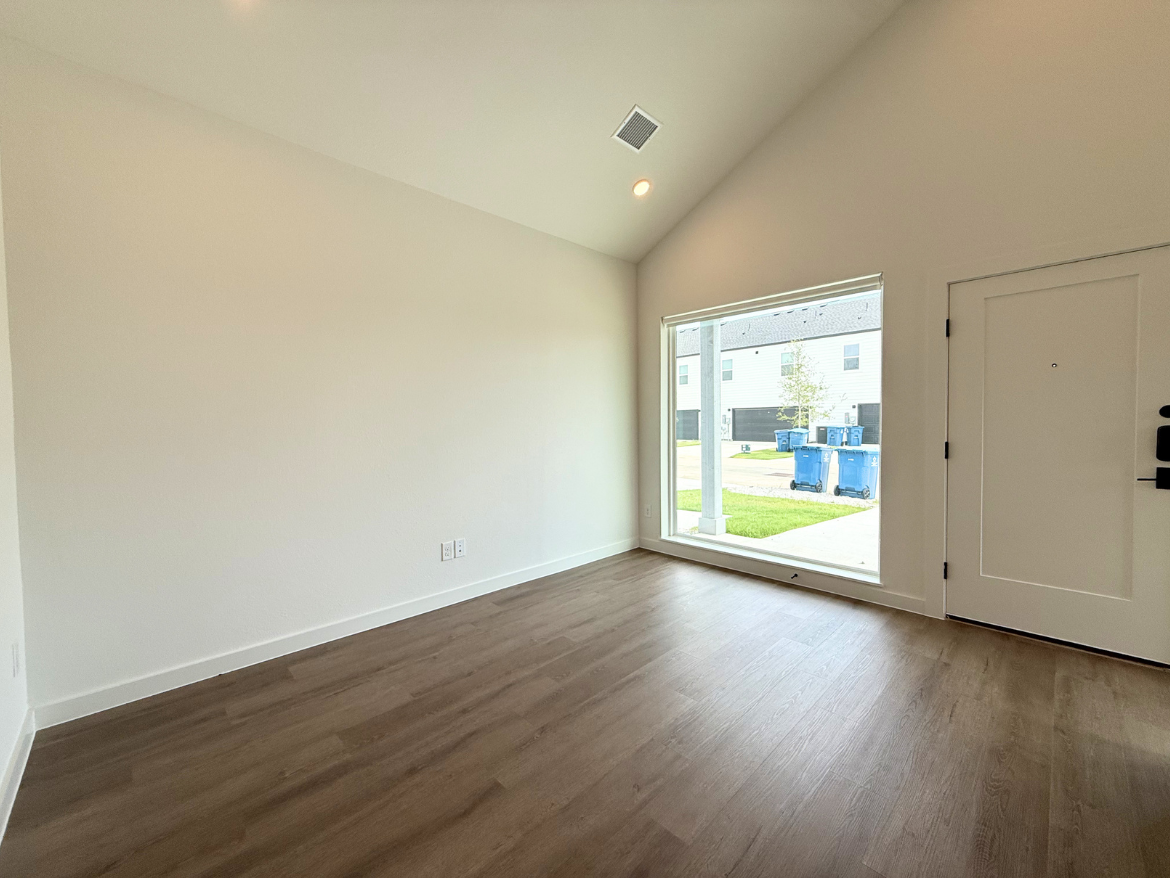 A spacious, empty room features light-colored walls and a high, sloped ceiling. The floor is made of light wood laminate. A large window on one wall allows natural light to flood the space, offering a view of a grassy area and blue trash bins outside. The entrance door is visible on the right side of the image. The overall atmosphere is bright and airy, with a minimalist design.