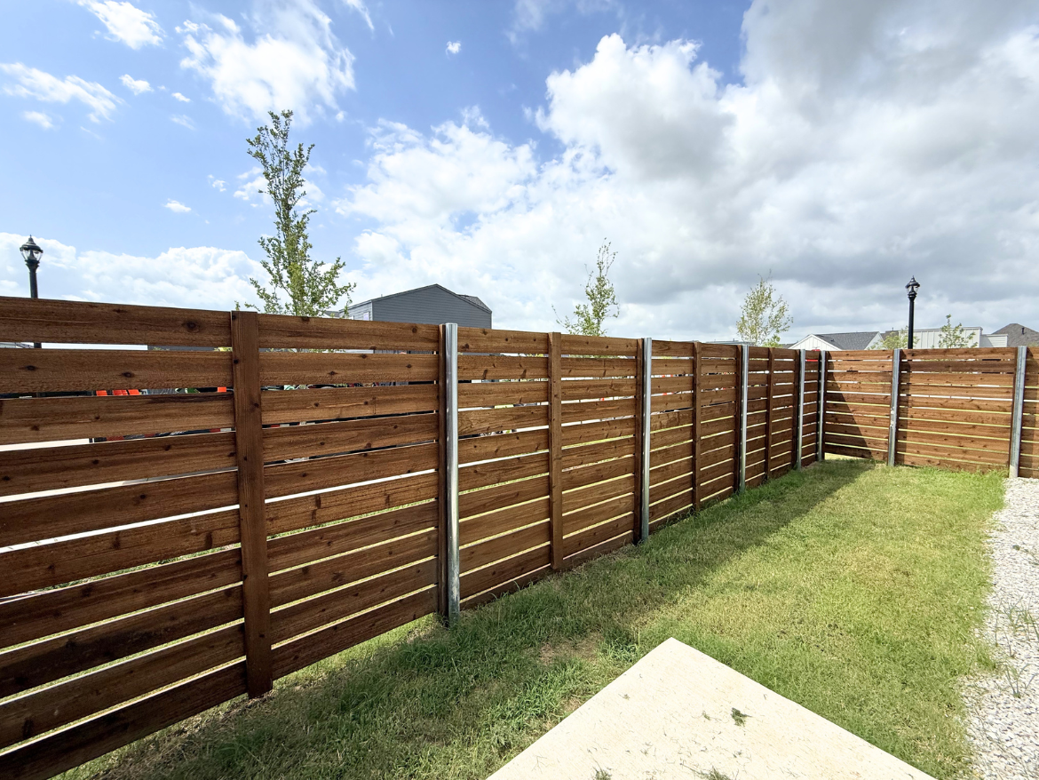 A wooden fence made of horizontal slats runs along the perimeter of a grassy area. The fence has a rich brown color and is supported by metal posts. In the background, there are a few small trees and a gray building. The sky is partly cloudy, with patches of blue visible. A light fixture can be seen in the distance, adding to the outdoor setting. The ground is a mix of grass and gravel.