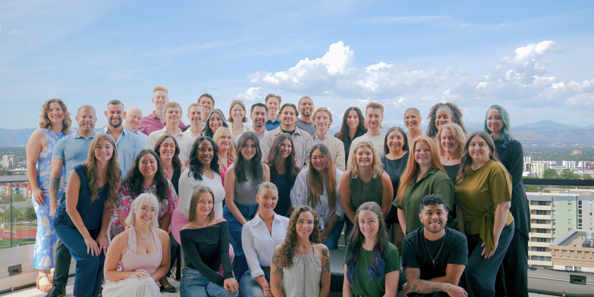 A large group of people in apartment locating are gathered outdoors, posing for a photo on a balcony with a scenic view. The group consists of both men and women, with a diverse range of hairstyles and clothing styles. They are smiling and appear to be enjoying the moment. The background features a clear blue sky with some clouds and distant mountains, along with urban buildings visible below. The atmosphere is cheerful and vibrant.
