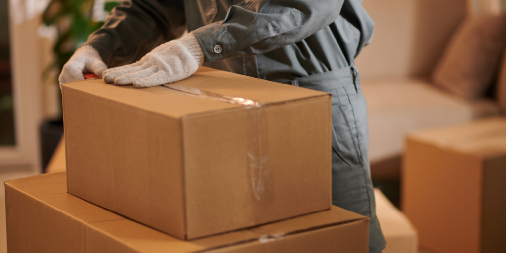 A person wearing gray gloves and a matching jumpsuit is placing a cardboard box on top of a stack of similar boxes. The boxes are sealed with tape and are situated in a room with soft furnishings in the background, suggesting a domestic or office environment. The setting appears warm and inviting, with greenery visible nearby. Moving in Austin, Texas.