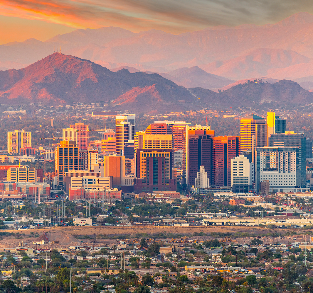 A panoramic view of a city skyline during sunset, showcasing a mix of modern skyscrapers and buildings. The foreground features a lower urban area with residential structures and greenery. In the background, rolling mountains are illuminated by the warm hues of the setting sun, creating a picturesque backdrop. The sky is painted with soft orange and pink tones, enhancing the overall beauty of the scene.
