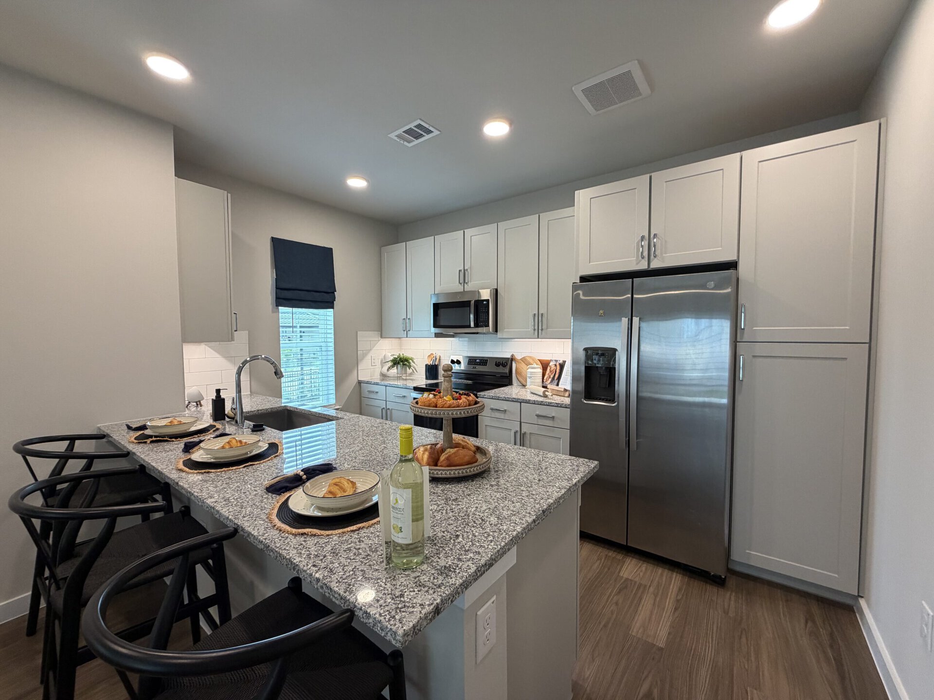 A modern kitchen featuring light gray cabinetry and stainless steel appliances. The countertop is made of gray speckled granite, with a circular tiered serving tray displaying pastries and fruit. Four black chairs are positioned around the island, which is set with plates, napkins, and a bottle of white wine. A window with a blue fabric shade allows natural light to enter, illuminating the space. The walls are painted in a soft neutral tone, enhancing the contemporary feel.