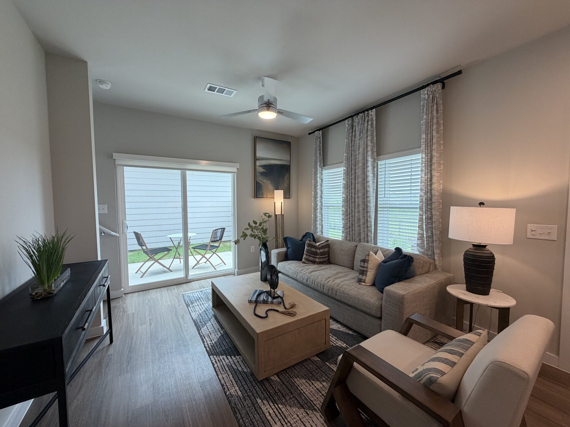 A modern living room features a light gray sofa adorned with various decorative pillows. In front of the sofa is a wooden coffee table with a few books and a decorative item on top. A floor lamp stands beside the sofa, providing soft lighting. Large windows with blinds allow natural light to fill the space, and sheer curtains add a touch of elegance. In the background, sliding glass doors lead to a small outdoor area with a table and two chairs, framed by a patch of green grass. A dark console table with a decorative plant completes the inviting atmosphere.