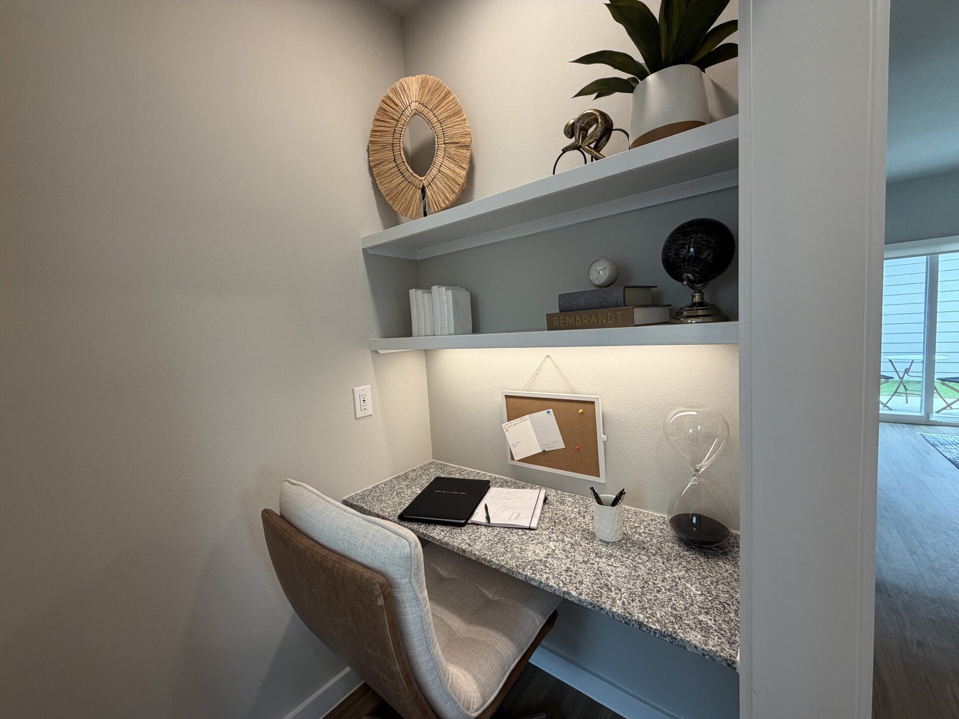 A cozy workspace features a granite desk with a light-colored chair. Above the desk, there are two shelves displaying decorative items, including a round mirror, a small globe, and books. The desk is organized with a black folder, a notepad, and a white cup holding pens. A corkboard with pinned notes hangs on the wall, and an hourglass sits on the desk. Natural light filters in from a nearby window, enhancing the inviting atmosphere.