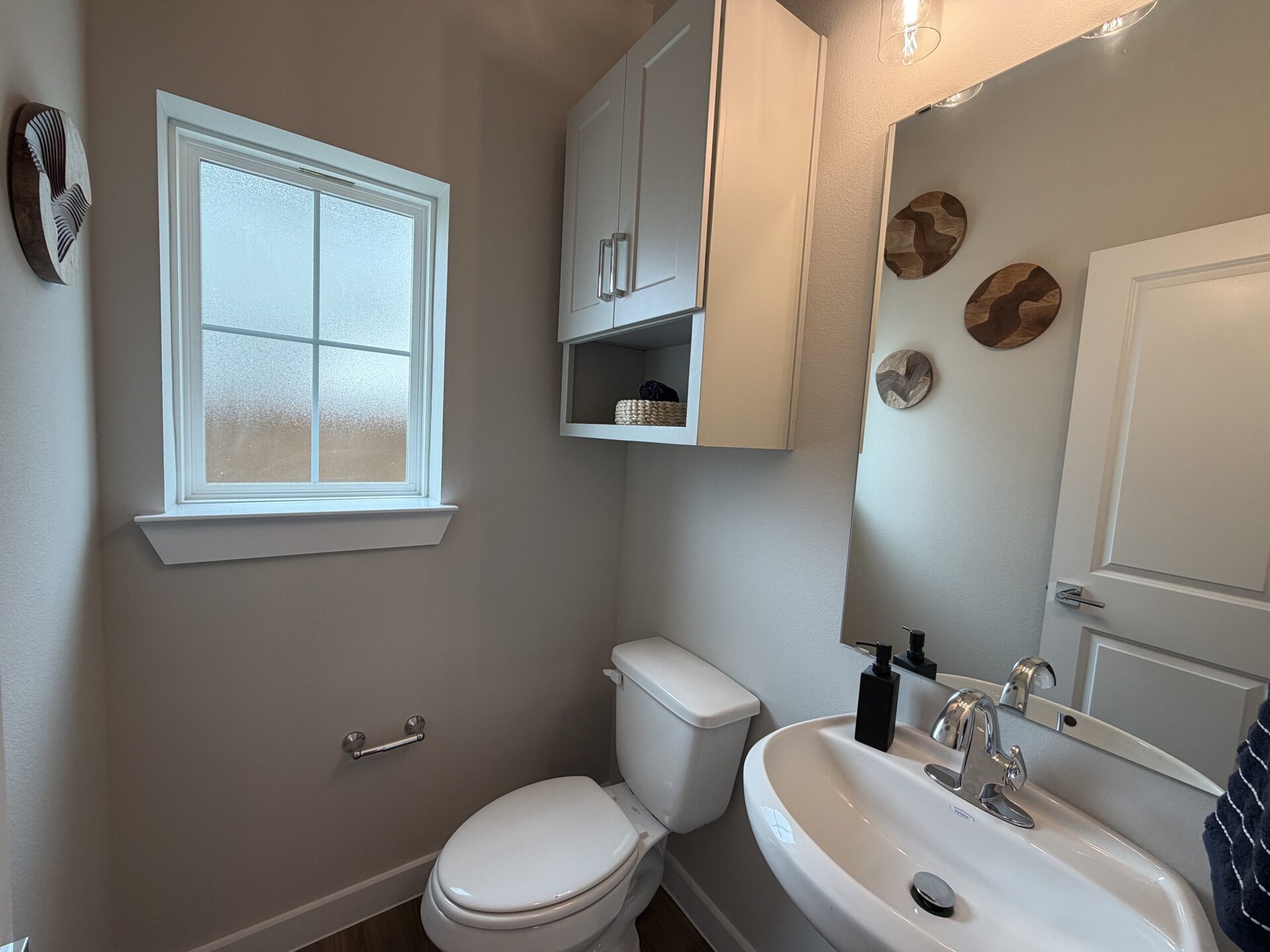 A small bathroom features a white toilet and a round sink with a chrome faucet. Above the sink, there is a mirror and a black soap dispenser. To the left, a window with frosted glass allows light in, while a small shelf above the toilet holds a woven basket. The walls are painted in a light color, and three wooden decorative pieces are mounted on the wall. A towel bar is positioned next to the toilet.