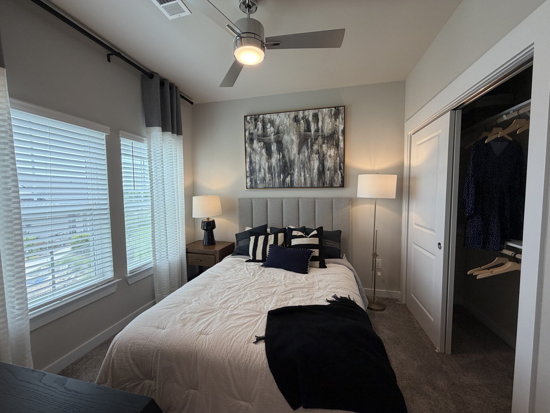 A cozy bedroom features a queen-sized bed with a light-colored quilt and an array of decorative pillows in navy and white. Above the bed hangs a large abstract painting in muted tones. To the side, a wooden nightstand holds a stylish black lamp. Natural light streams in through two large windows adorned with sheer white curtains, while a floor lamp stands beside the bed. A closet with sliding doors is visible, showcasing a dress on a hanger. The room has a neutral color palette and a soft carpeted floor.