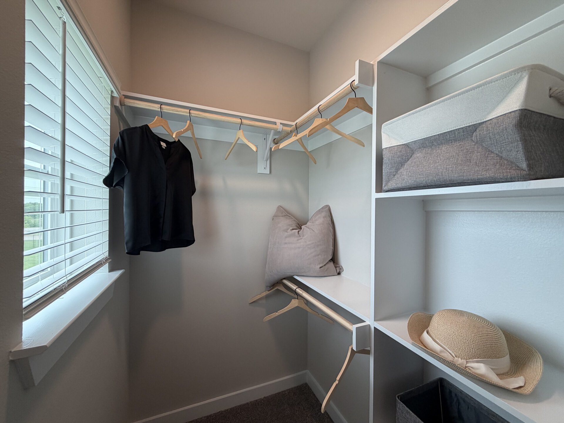 A small closet features a light gray wall and a window with blinds. A black short-sleeve shirt hangs on a wooden hanger from a rod above. Below, there are additional wooden hangers and a plush gray pillow resting on a shelf. On another shelf, a gray and white storage box sits beside a straw hat. The overall design is clean and organized, with a focus on functionality.