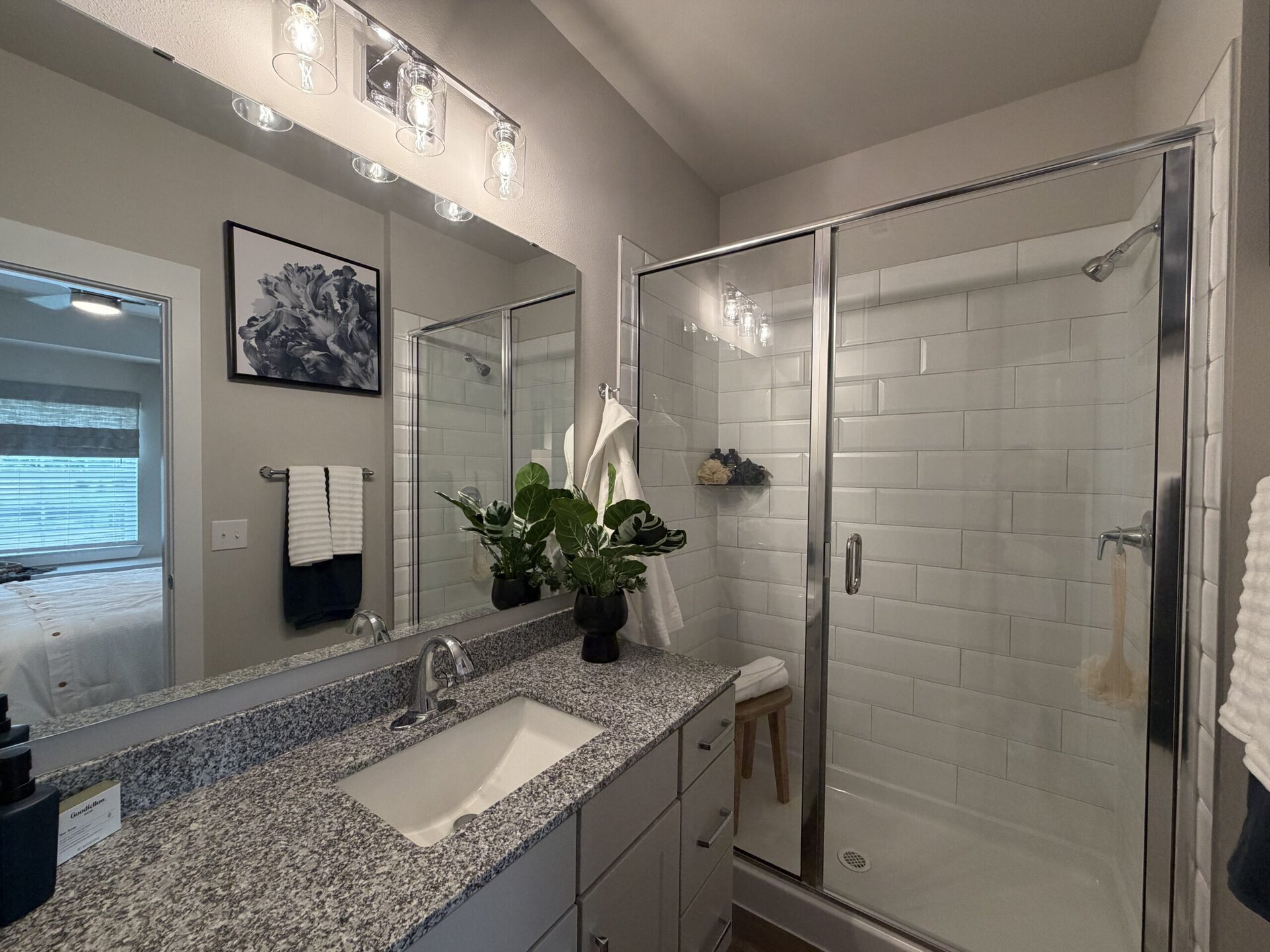 A modern bathroom features a glass shower enclosure with white subway tiles. A granite countertop with an undermount sink is complemented by a sleek faucet. Above the sink, a large mirror reflects the space, which includes a towel rack with neatly folded towels. A potted plant adds a touch of greenery on the counter. The lighting consists of stylish glass fixtures, and a piece of artwork hangs on the wall. In the background, a glimpse of a bedroom with a window and blinds is visible.