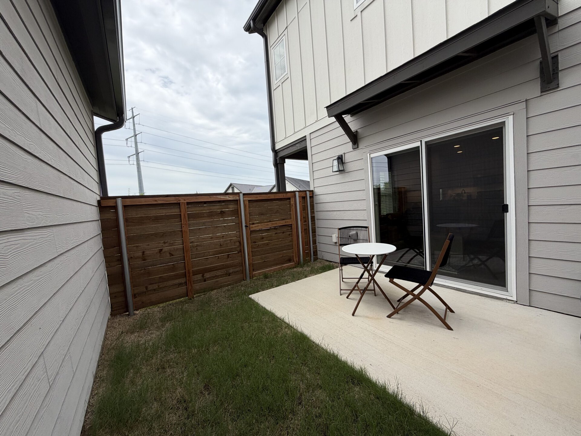 A small outdoor patio area features a round white table and two folding chairs. The patio is surrounded by a wooden fence, with grass growing nearby. In the background, a portion of a house is visible, along with a sliding glass door. Power lines can be seen in the distance against a cloudy sky.