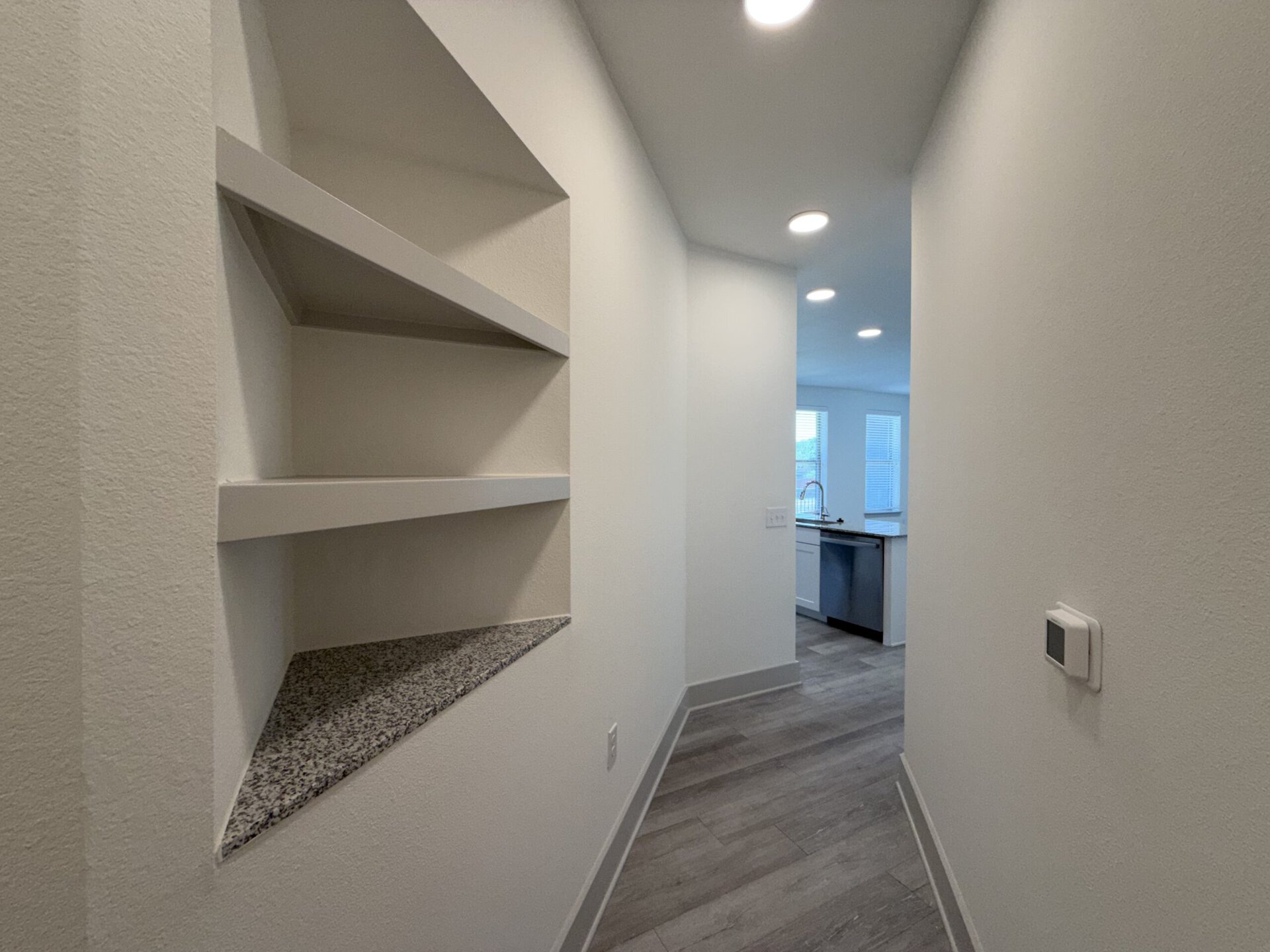 A narrow hallway features a built-in shelving unit with three white shelves, set against a light-colored wall. The floor is covered in light wood-like planks. At the end of the hallway, a kitchen area is visible, showcasing modern appliances and a sink, with large windows allowing natural light to enter. A small control panel is mounted on the wall to the right.