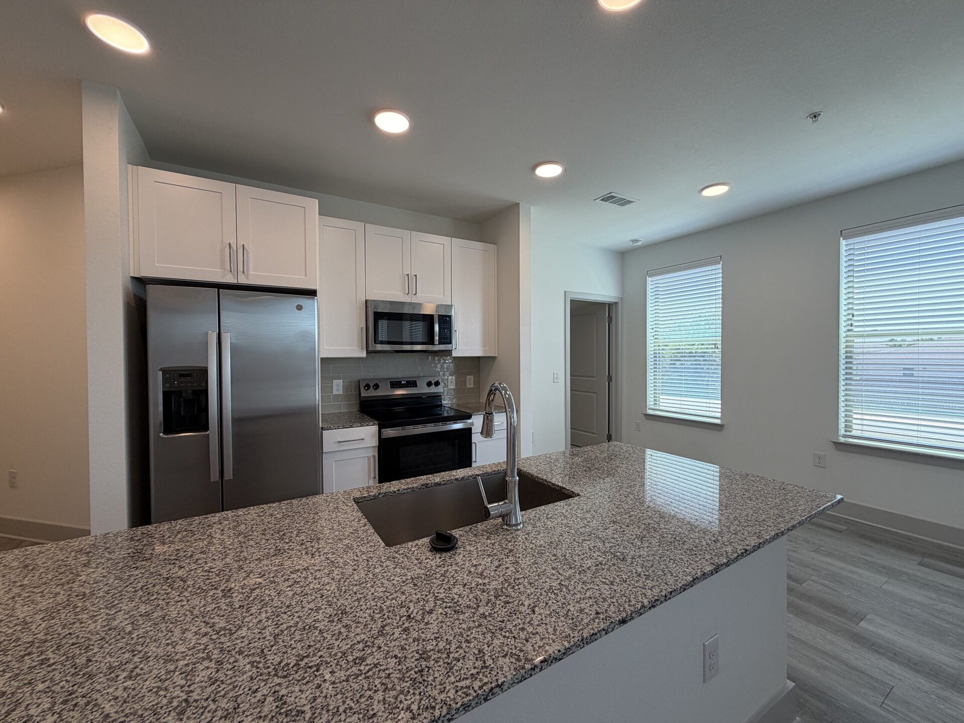 A modern kitchen features white cabinetry and stainless steel appliances, including a refrigerator, microwave, and oven. The countertop is made of speckled granite, with a sink integrated into the island. Natural light streams in through two windows with blinds, illuminating the space. The flooring is a light-colored tile, enhancing the contemporary feel of the kitchen.