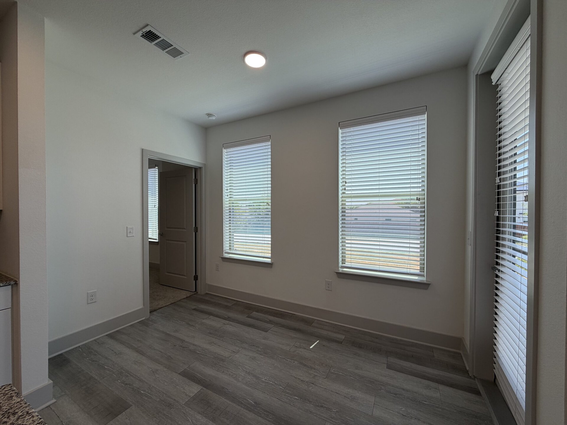A well-lit room features two windows with horizontal blinds, allowing natural light to enter. The walls are painted in a light color, and the flooring is a modern gray laminate. A door on the left leads to another space, while the overall design is clean and minimalistic, creating an inviting atmosphere.