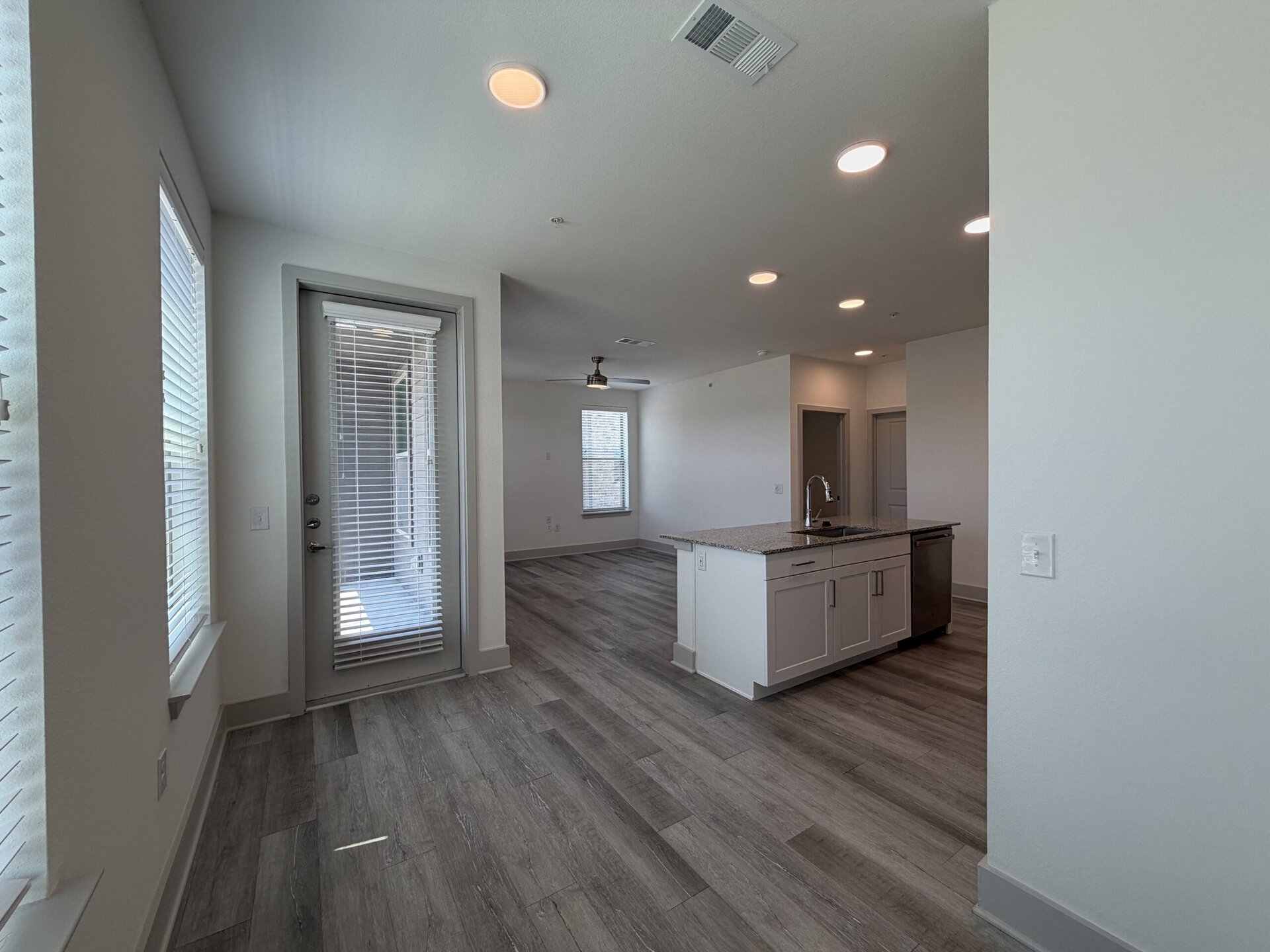 A spacious, modern interior features an open layout with light-colored walls and wood-like flooring. On the left, a door with blinds leads to an outdoor area, while large windows allow natural light to fill the space. The kitchen island, equipped with a sink, is positioned in the center, complemented by cabinetry and a stainless steel dishwasher. Recessed lighting illuminates the area, and a ceiling fan is visible in the living space. Doorways lead to additional rooms, enhancing the overall functionality of the layout.