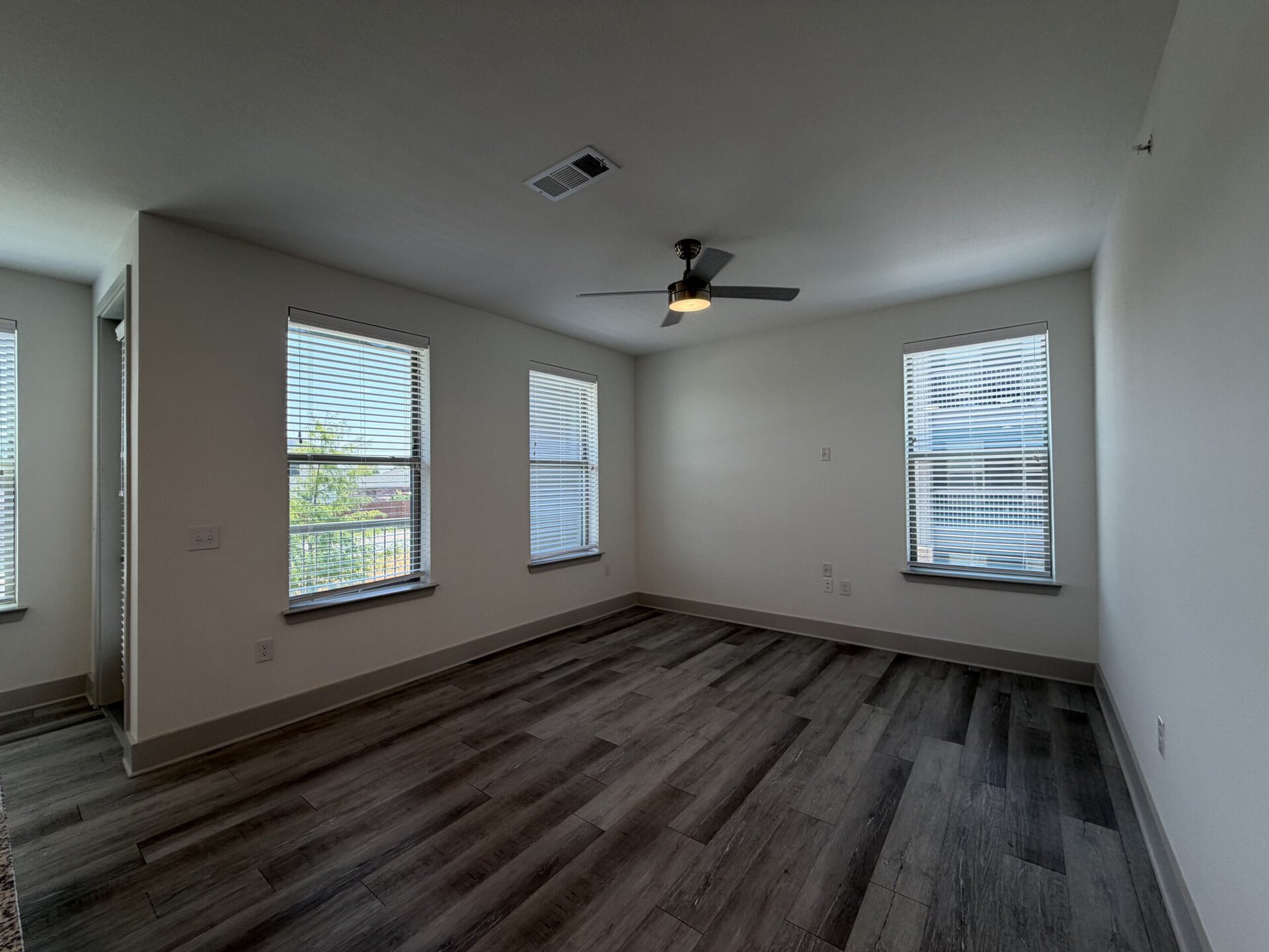 A bright, empty room features two windows with blinds, allowing natural light to enter. The walls are painted a light color, and the flooring consists of wood-like planks in a gray tone. A ceiling fan with a light fixture is mounted in the center of the room. There is a door on one side, leading to another space. The overall atmosphere is clean and modern.