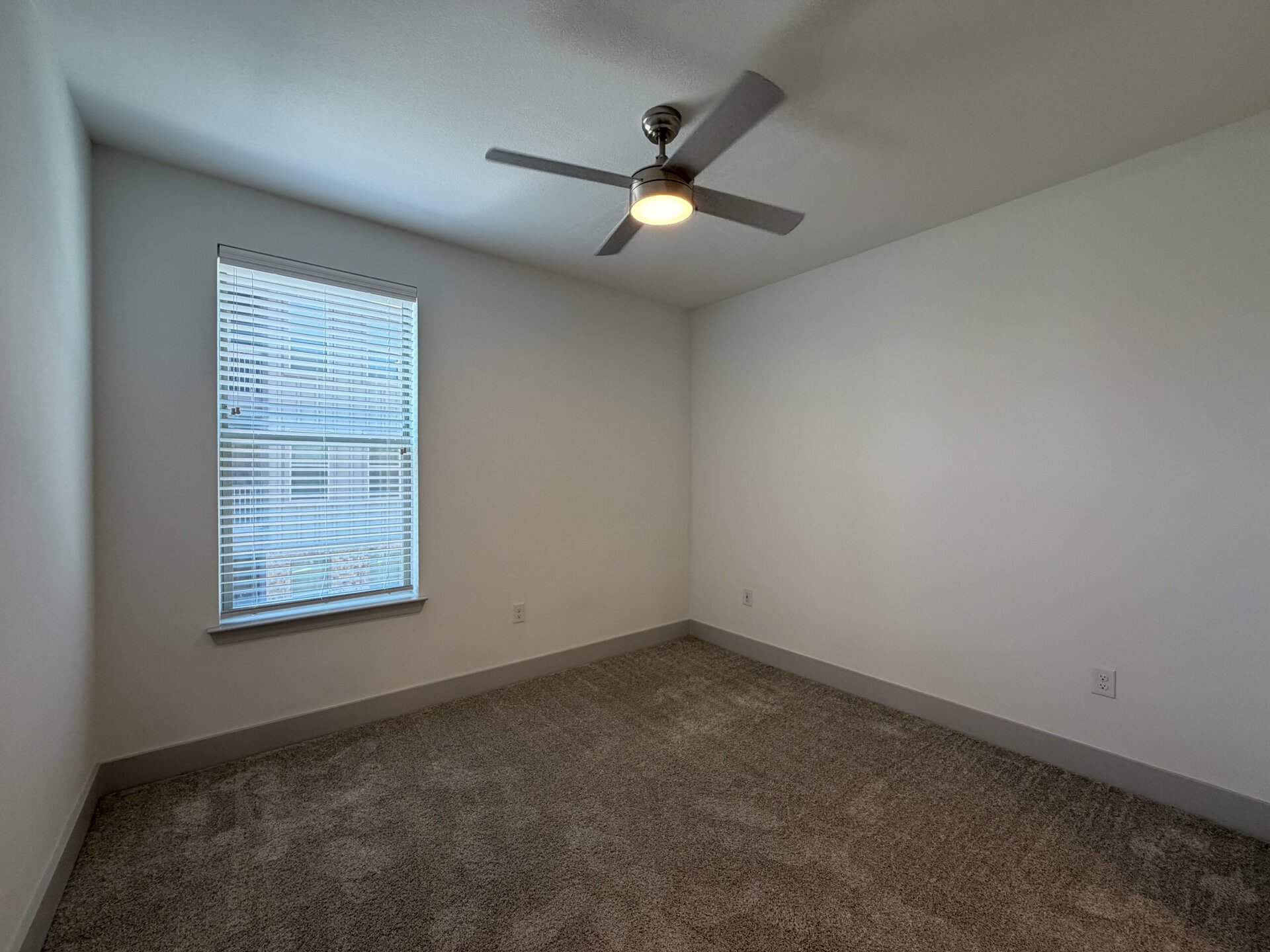 A small, empty room features light-colored walls and a soft, carpeted floor. A window with horizontal blinds allows natural light to enter, and a ceiling fan with a light fixture is mounted in the center of the ceiling. The room has a clean and minimalist appearance, with no furniture or decorations present.