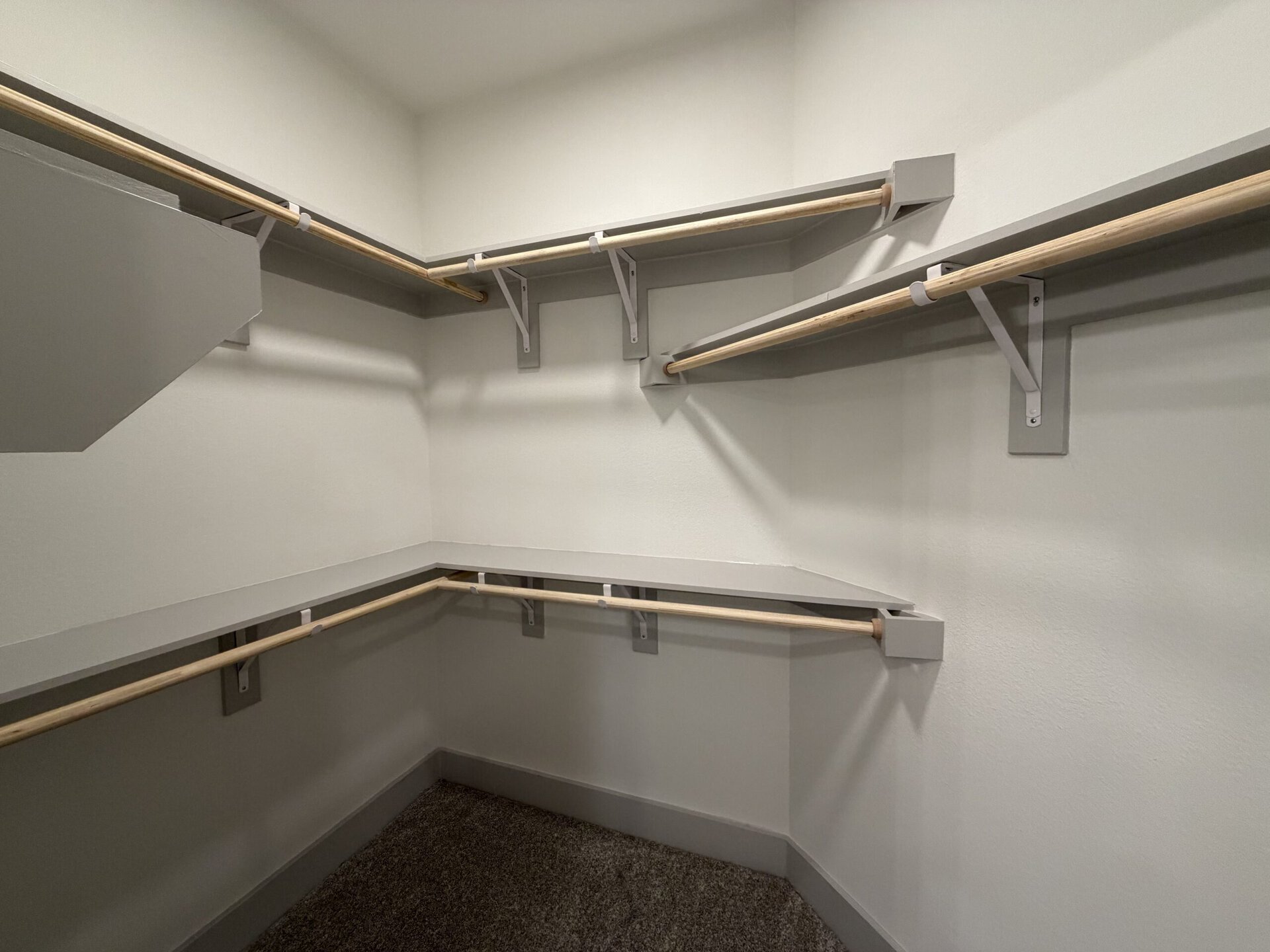 A spacious closet features light gray walls and shelving. There are multiple wooden rods for hanging clothes, supported by gray brackets. A long shelf runs along the back wall, providing additional storage space. The floor is carpeted in a neutral tone, enhancing the overall clean and organized appearance of the closet.