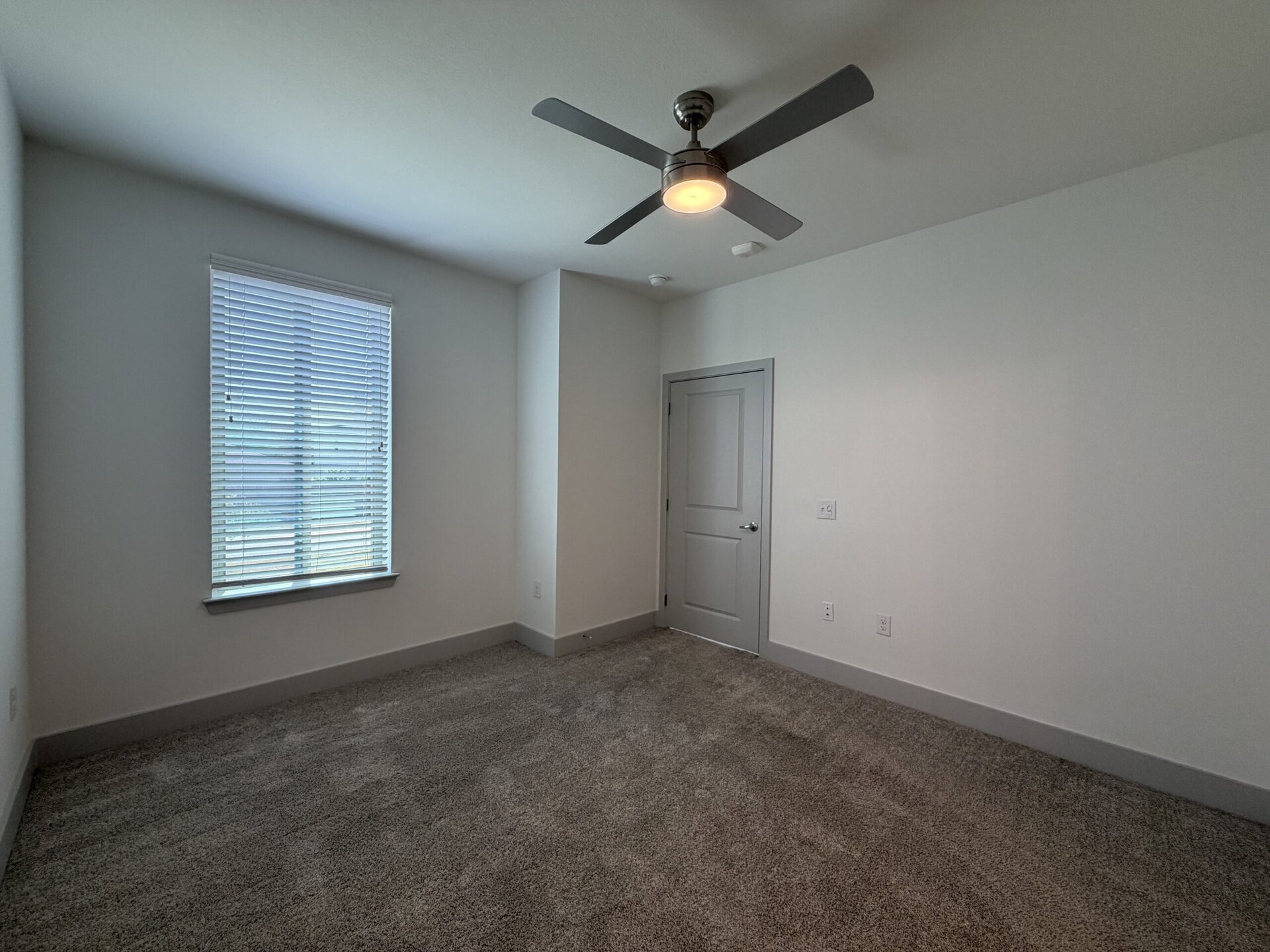 A spacious, empty room features light gray walls and a plush, light-colored carpet. A window with horizontal blinds allows natural light to enter, and a ceiling fan with a light fixture hangs above. A closed door is visible on the right side, leading to another space. The overall ambiance is bright and minimalistic.