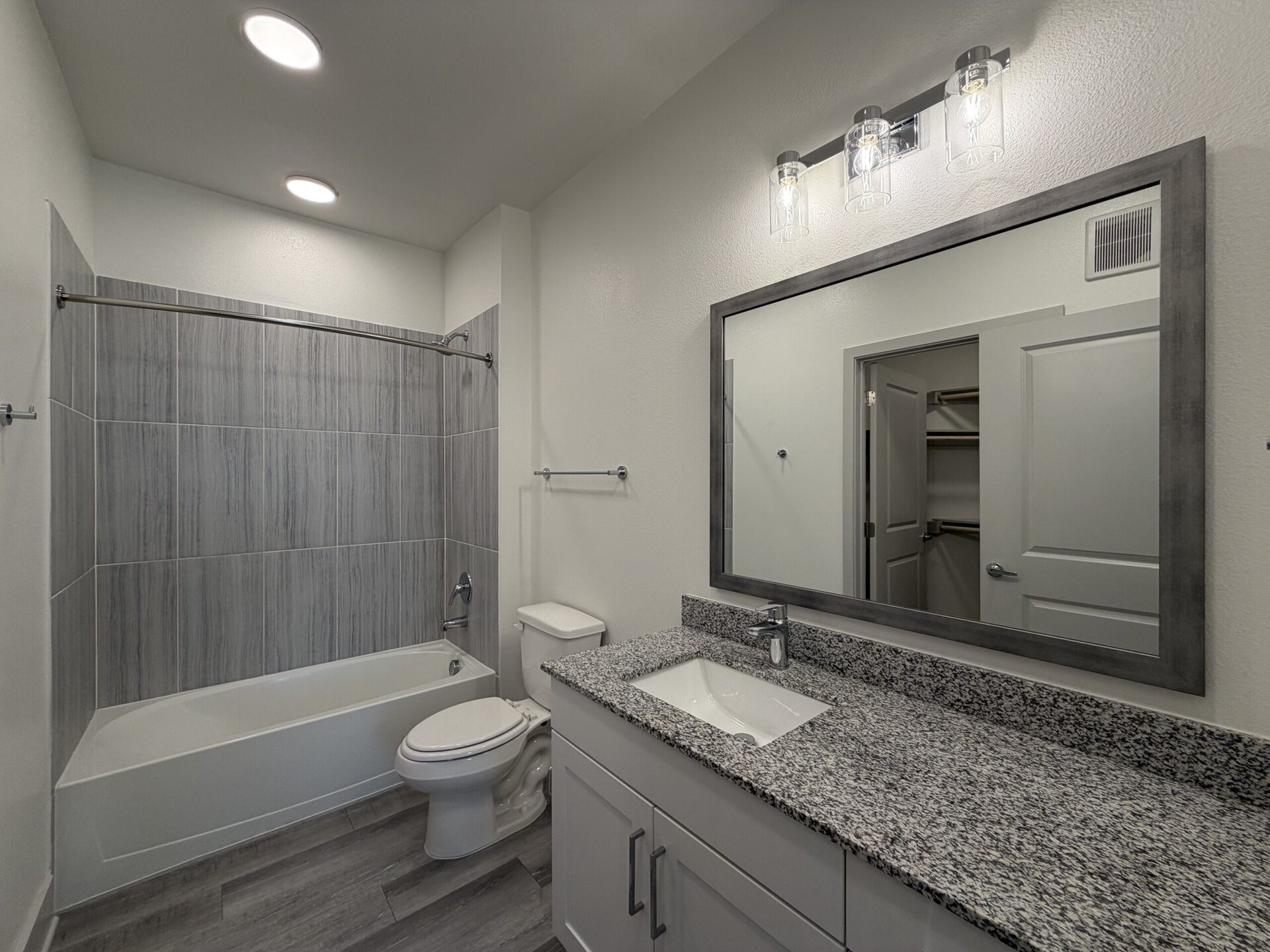 A modern bathroom features a shower-tub combo with gray tiled walls and a white tub. A white toilet is positioned next to the tub. The vanity has a granite countertop with a rectangular sink and a sleek faucet. Above the sink, a large mirror with a gray frame reflects the space. Three glass light fixtures hang above the mirror, providing illumination. A door leads to a closet area, visible in the background. The walls are painted a light color, enhancing the overall brightness of the room.