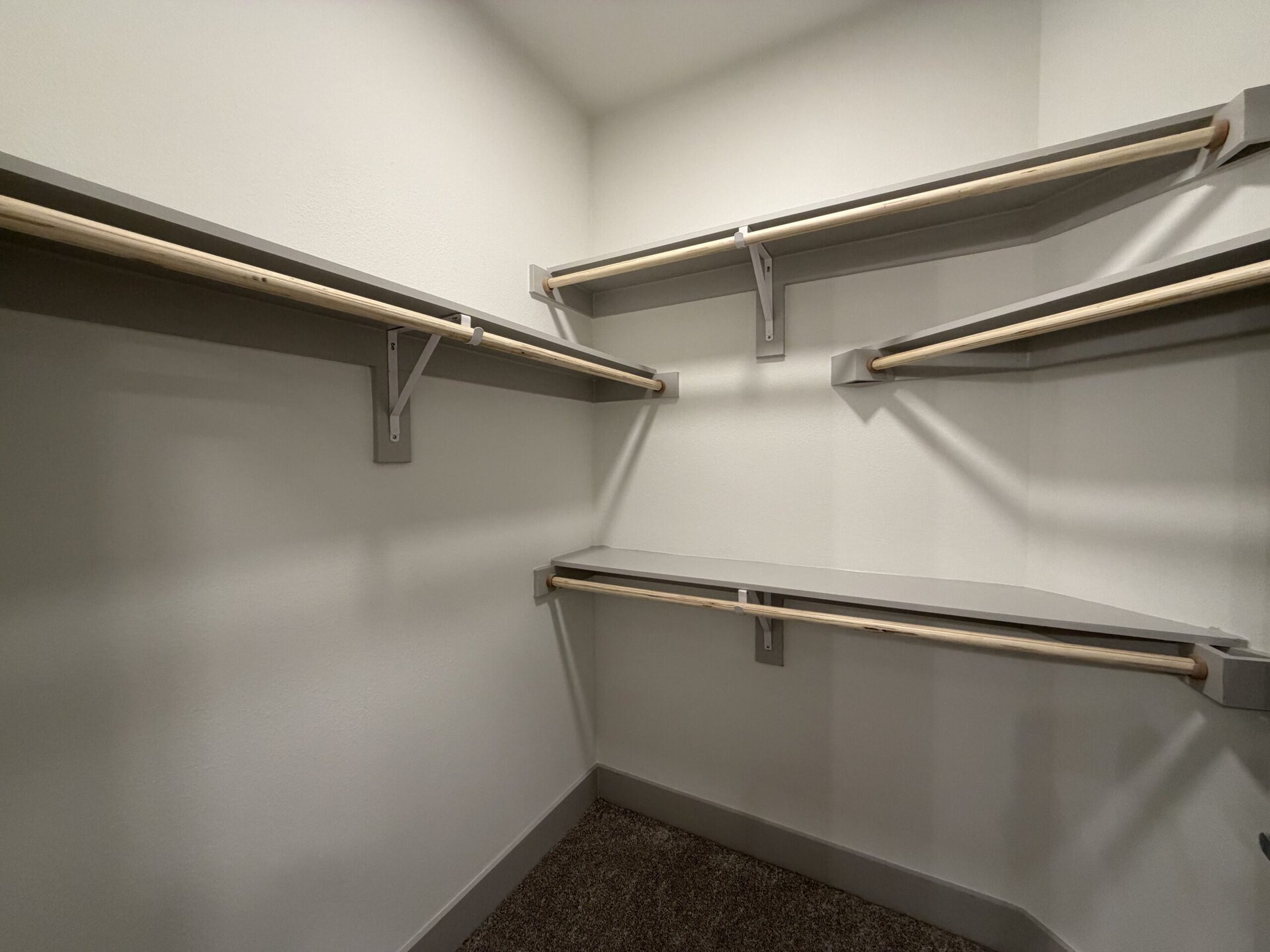 A spacious closet features light gray walls and a carpeted floor. It has multiple wooden rods for hanging clothes, supported by gray brackets. There are also two gray shelves, one on the left and one on the right, providing additional storage space. The overall design is simple and functional, with ample room for organization.
