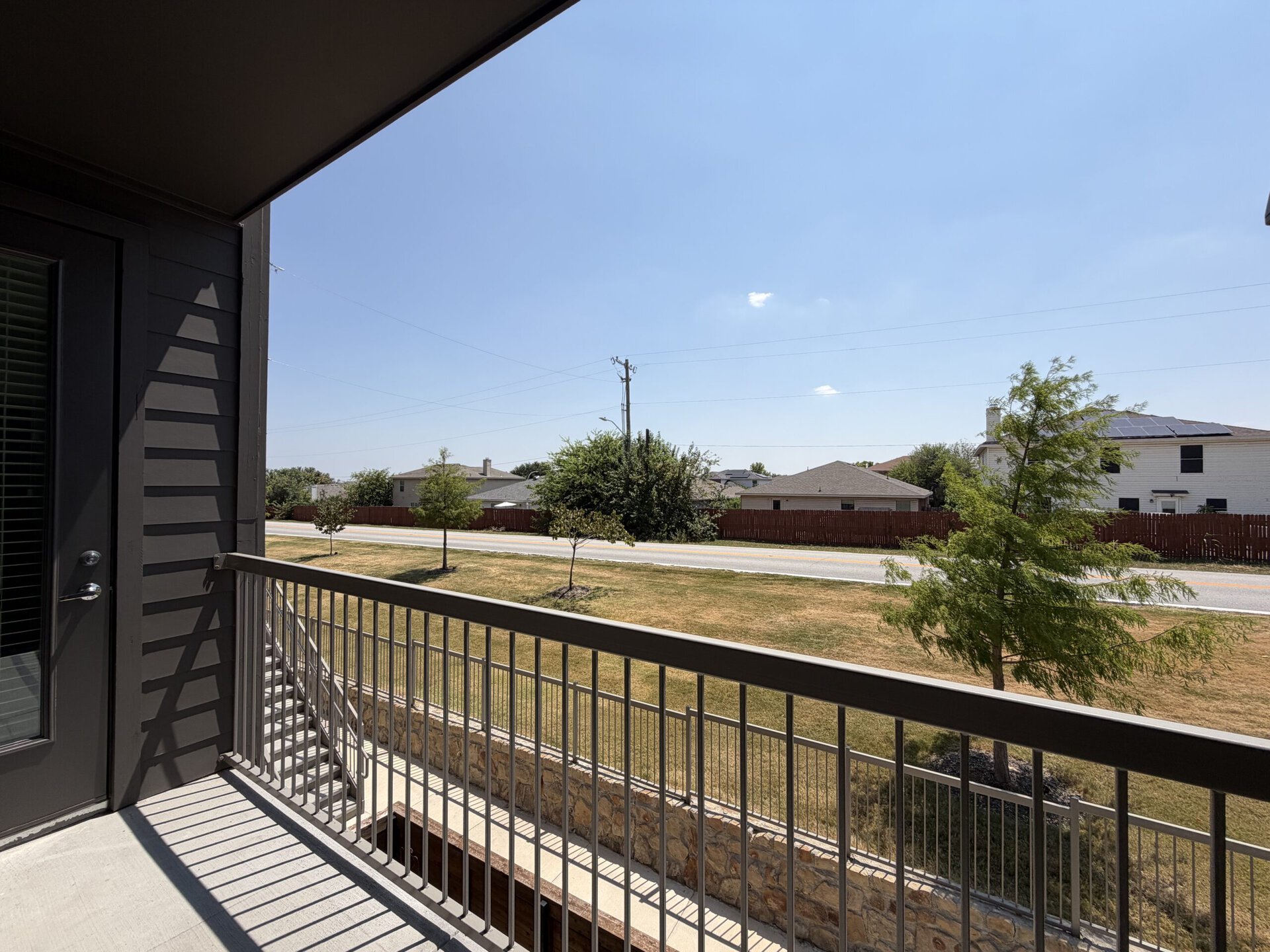 A view from a balcony shows a clear blue sky with a few clouds. In the foreground, a metal railing frames the scene. Below, a grassy area is visible, with a couple of small trees. Across the street, several houses can be seen, some with solar panels on their roofs, and a wooden fence borders the properties. The overall atmosphere is bright and sunny.