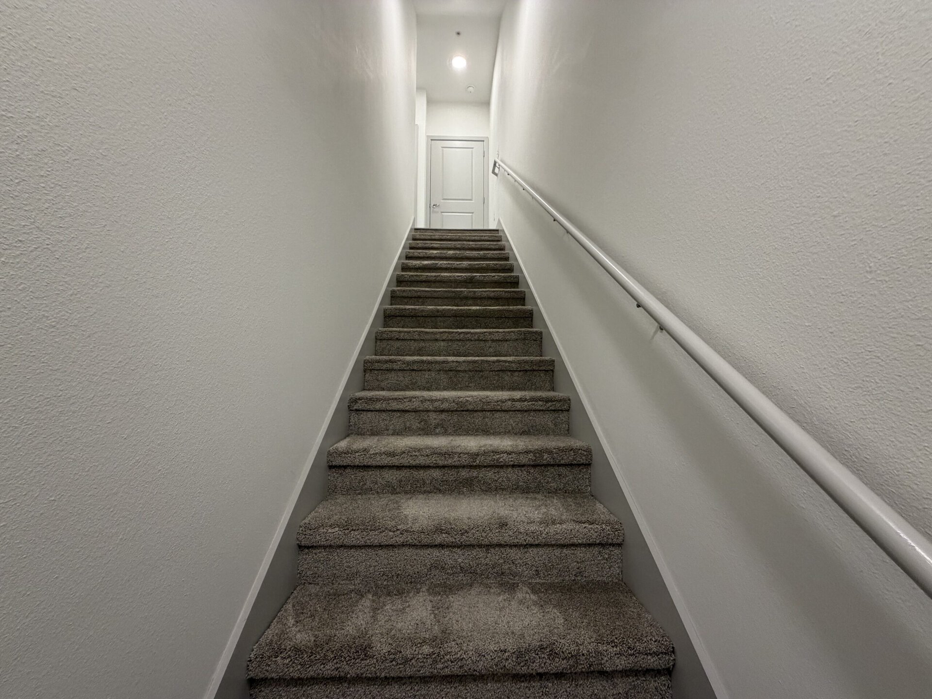A staircase with carpeted steps leads upward, flanked by plain white walls. A handrail runs along one side. At the top of the stairs, a closed white door is visible, illuminated by soft lighting. The overall color scheme is neutral, creating a clean and modern appearance.